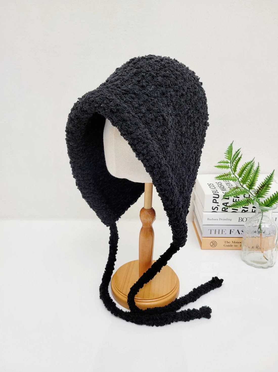 Black sherpa little riding hood bonnet hat on a wooden stand, featuring long, soft ties.