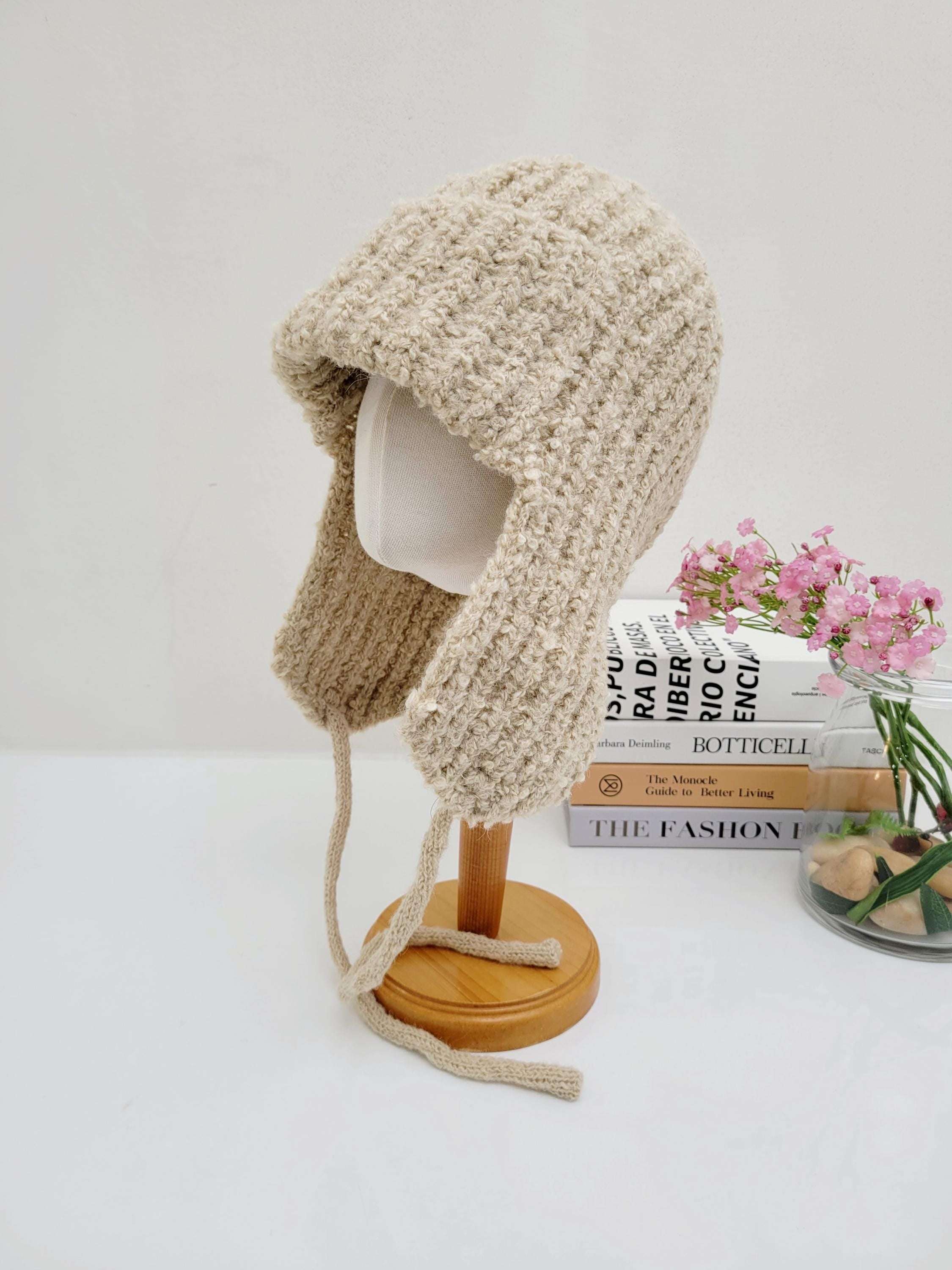 Beige knitted wool earflap hat on a stand. Features soft texture, ear ties, and cozy design, perfect for winter warmth.