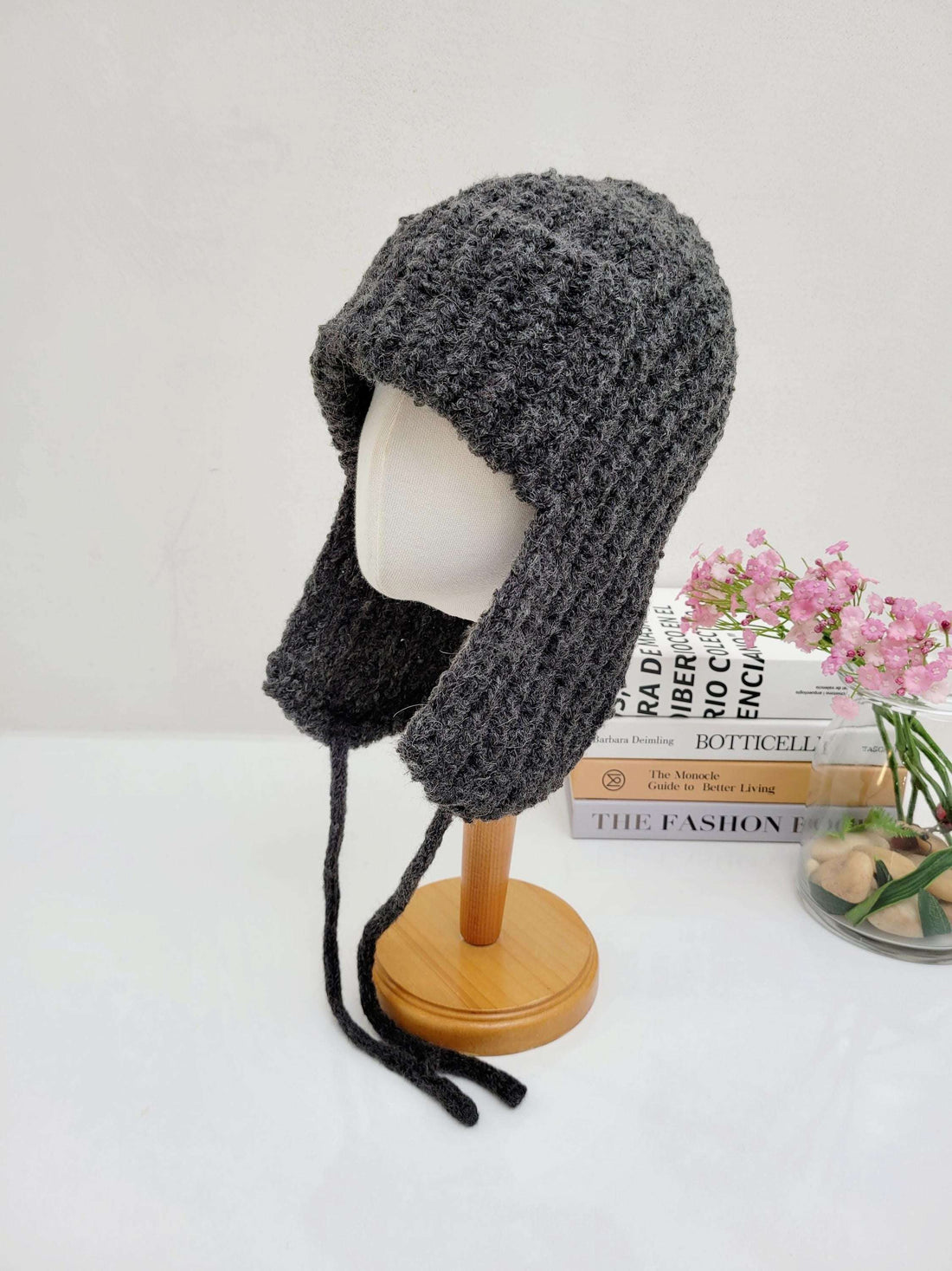 Gray knitted wool earflap hat on a stand, featuring long tie strings and a textured knit pattern.