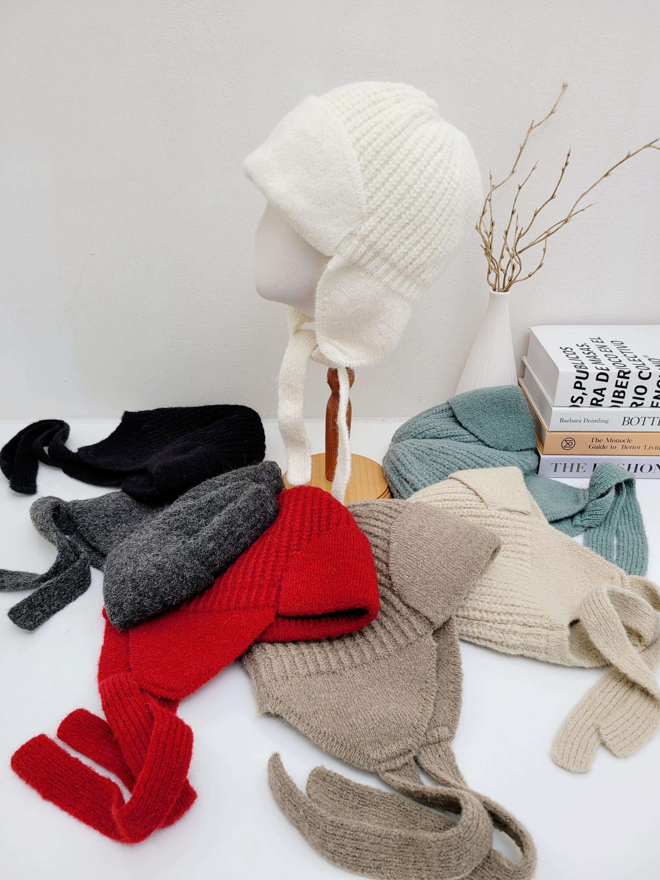 Assorted amelia knit hats in white, black, gray, red, beige, and teal. Each hat features ear flaps with ties, displayed on a white surface with books and a vase.