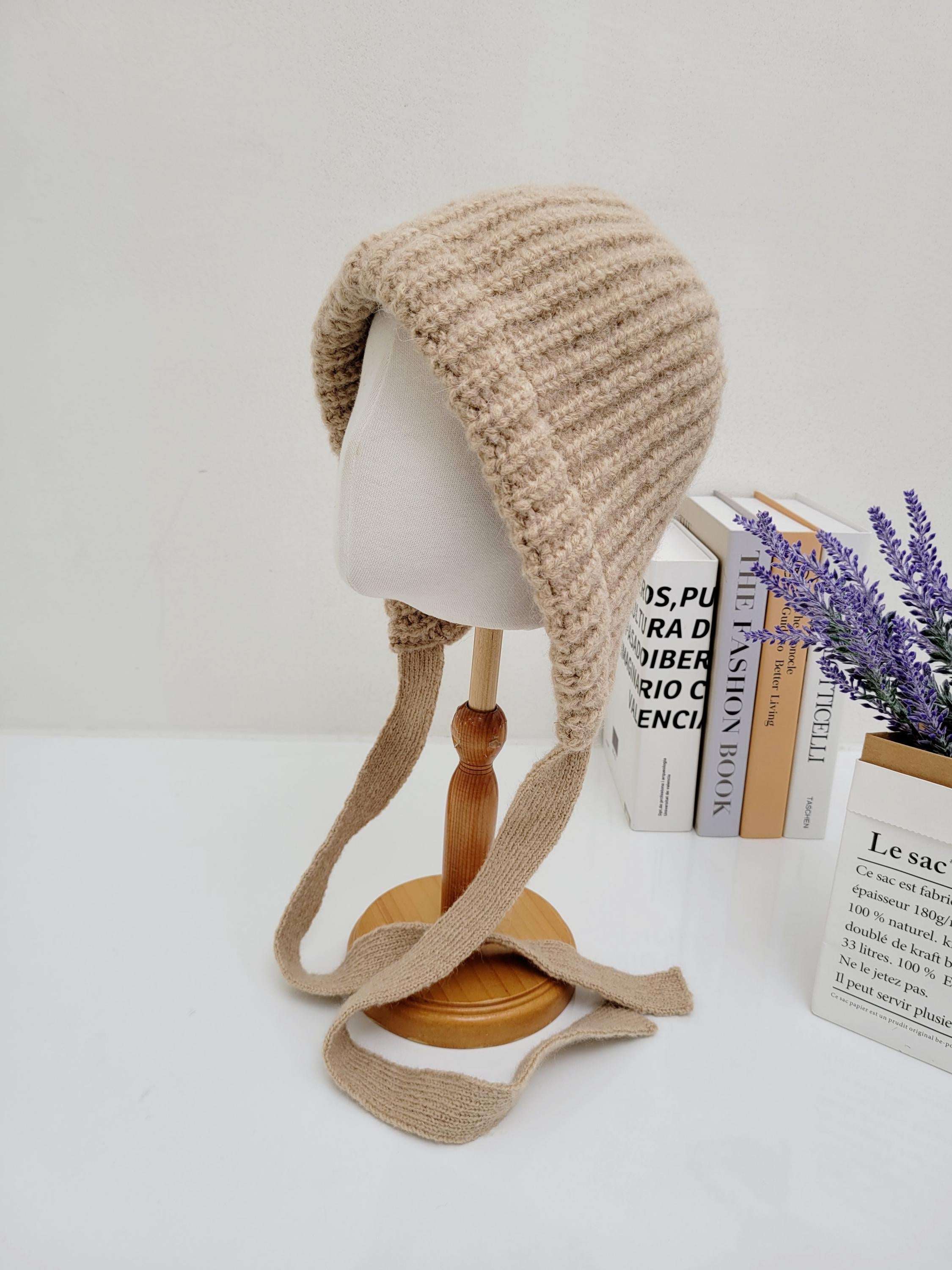 Beige knit tour le jour hat with ear flaps and tie strings, displayed on a mannequin head.