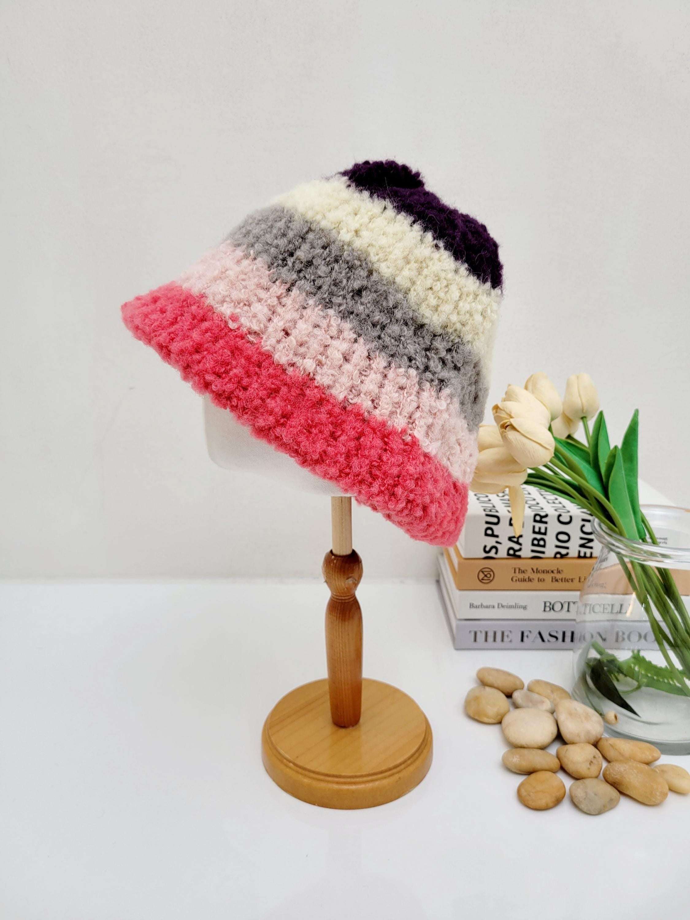 Striped tokyo beanie in pink, grey, cream, and purple knit. A cozy and stylish hat on a wooden display stand.