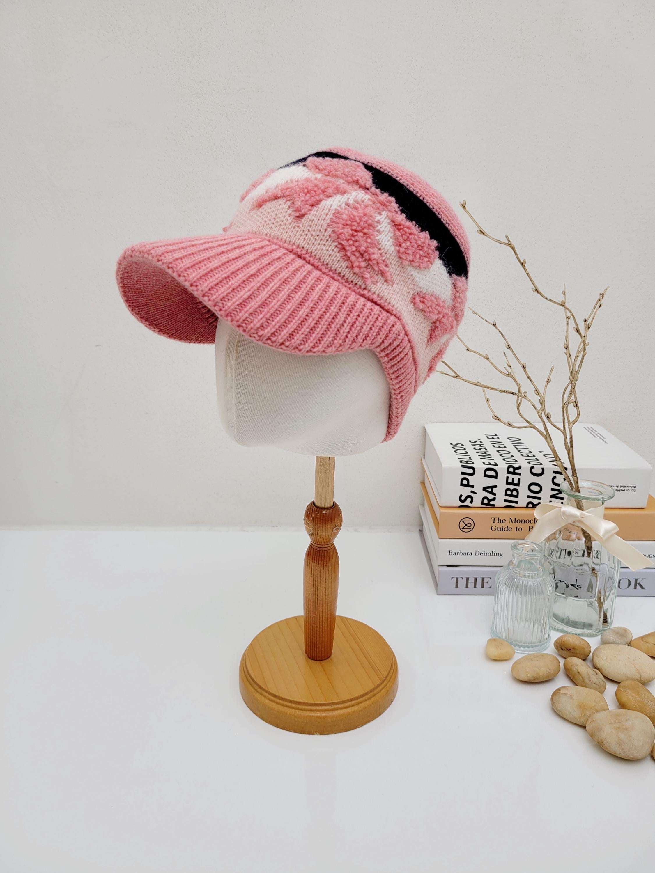 Pink earmuff knitted wool cap with a visor and black/white patterned accents, displayed on a wooden stand.