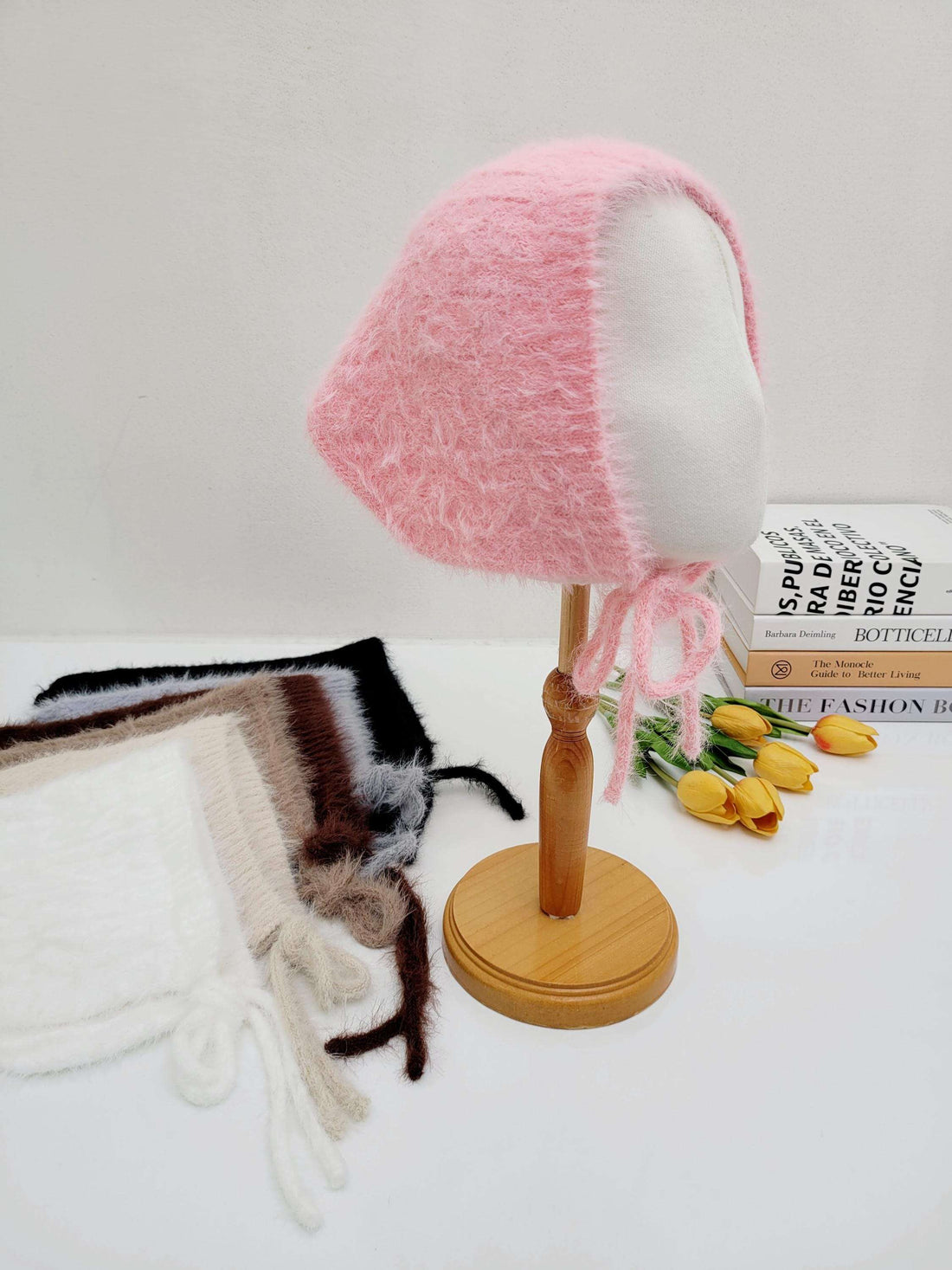 Pink fuzzy little riding hood bonnet on display; soft texture, with tie strings. Other bonnets in white, gray, brown and black are visible.