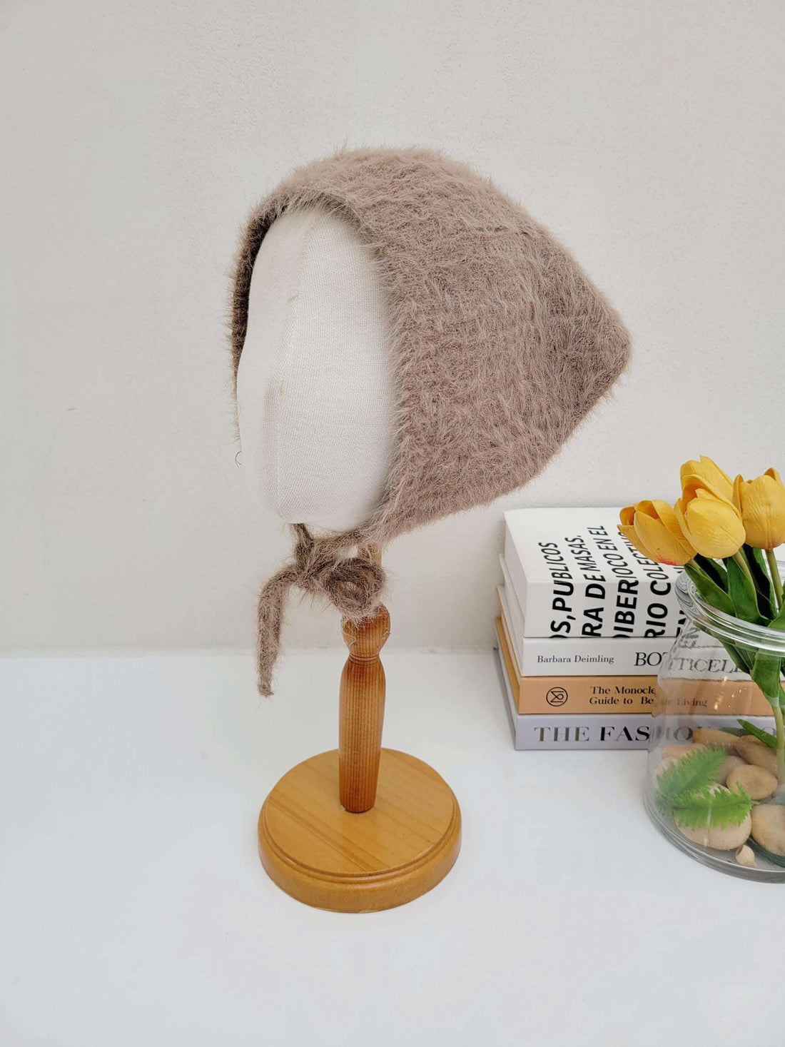 Brown, fuzzy "little riding hood" bonnet tied under the chin, displayed on a mannequin head. Cozy and stylish headwear.