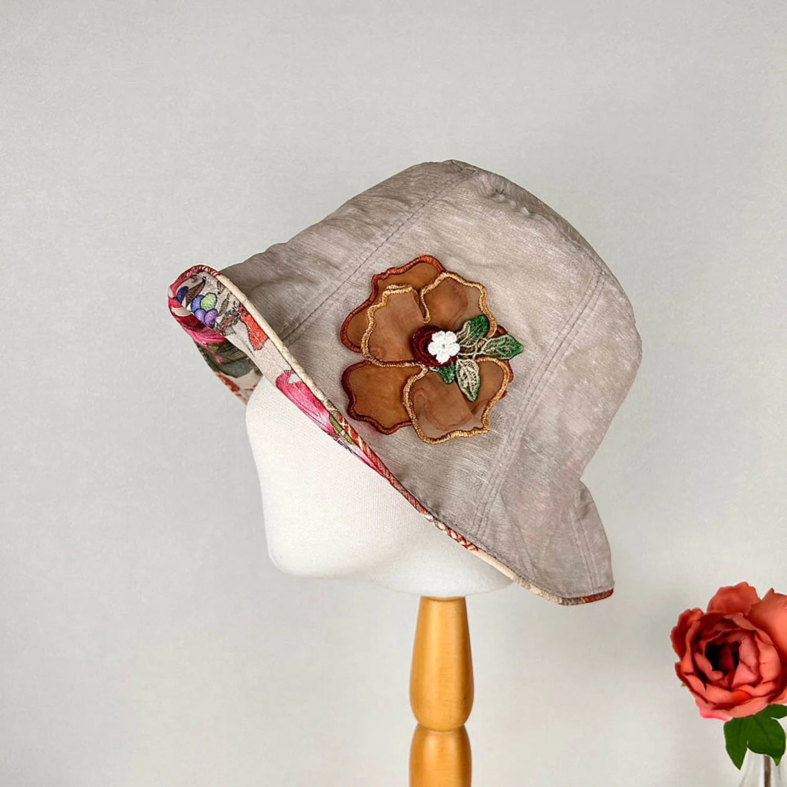 Beige linen bucket-side flower hat with brown floral appliqué and colorful floral-print brim, displayed on a mannequin.