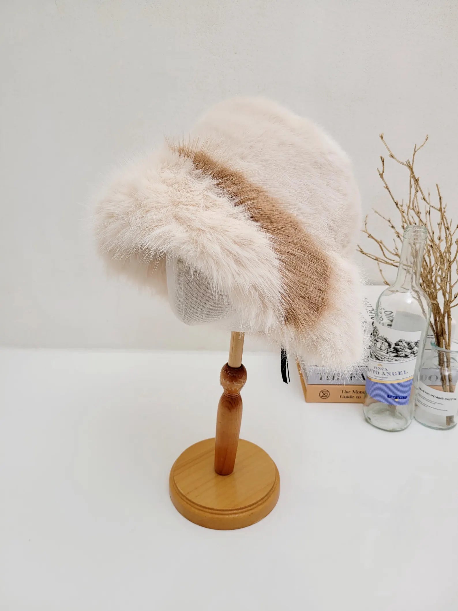 Creamy white fluffy vegan fur hat with a brown stripe, displayed on a wooden stand, perfect for winter warmth and style