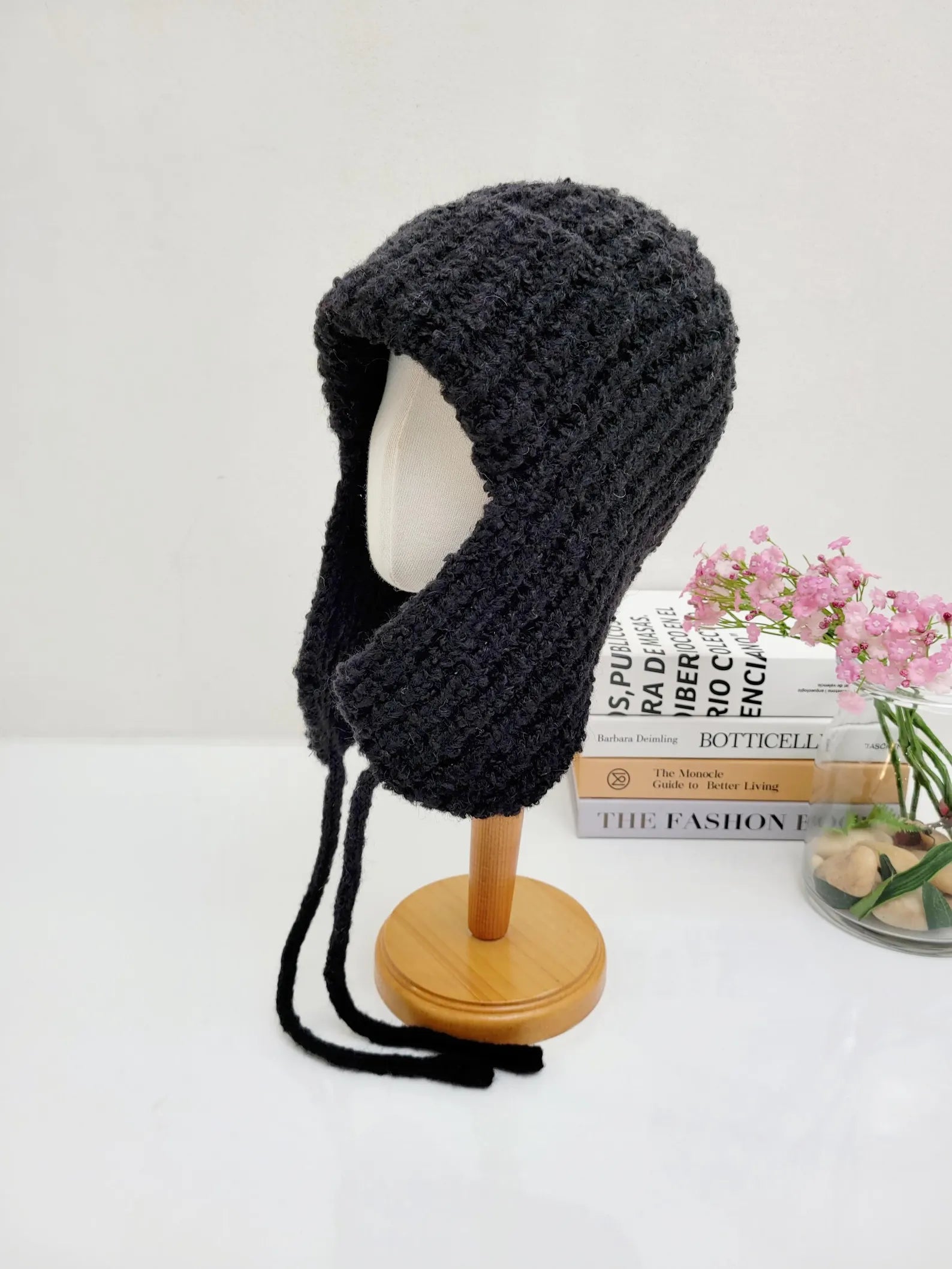 Black knitted wool earflap hat with cozy texture and long tie strings, displayed on a stand.