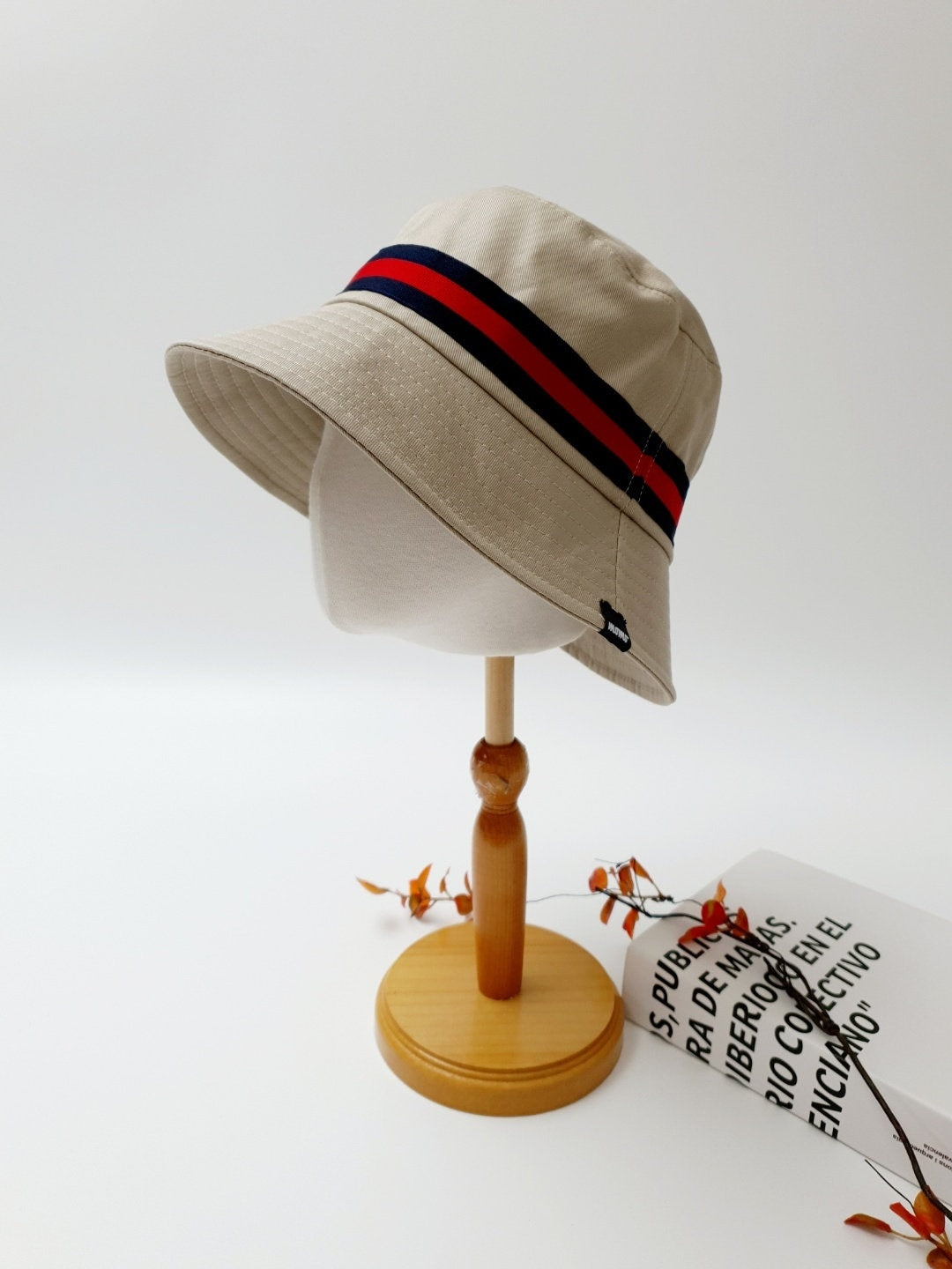 Beige daytona bucket hat with red and navy band, displayed on a wooden stand. Casual style with a touch of nautical flair.