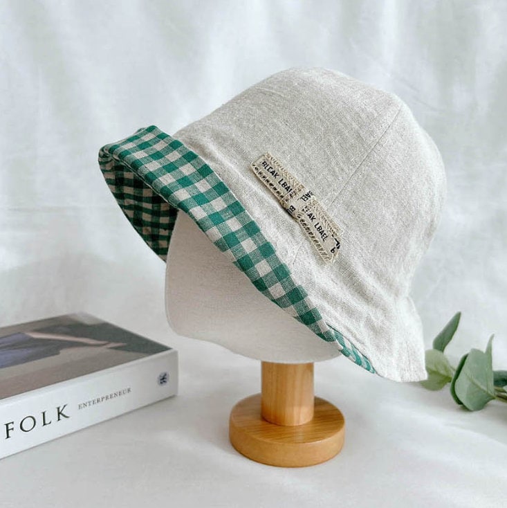 Beige linen checkered bucket hat with a green and white gingham brim, displayed on a wooden stand.