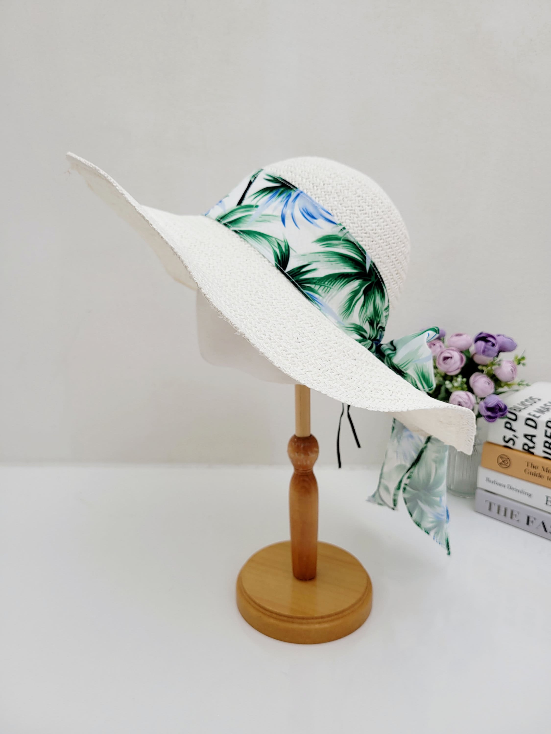 EAST HAMPTON: White sun hat with a wide brim, accented by a vibrant tropical print ribbon with green palm leaves and blue accents.