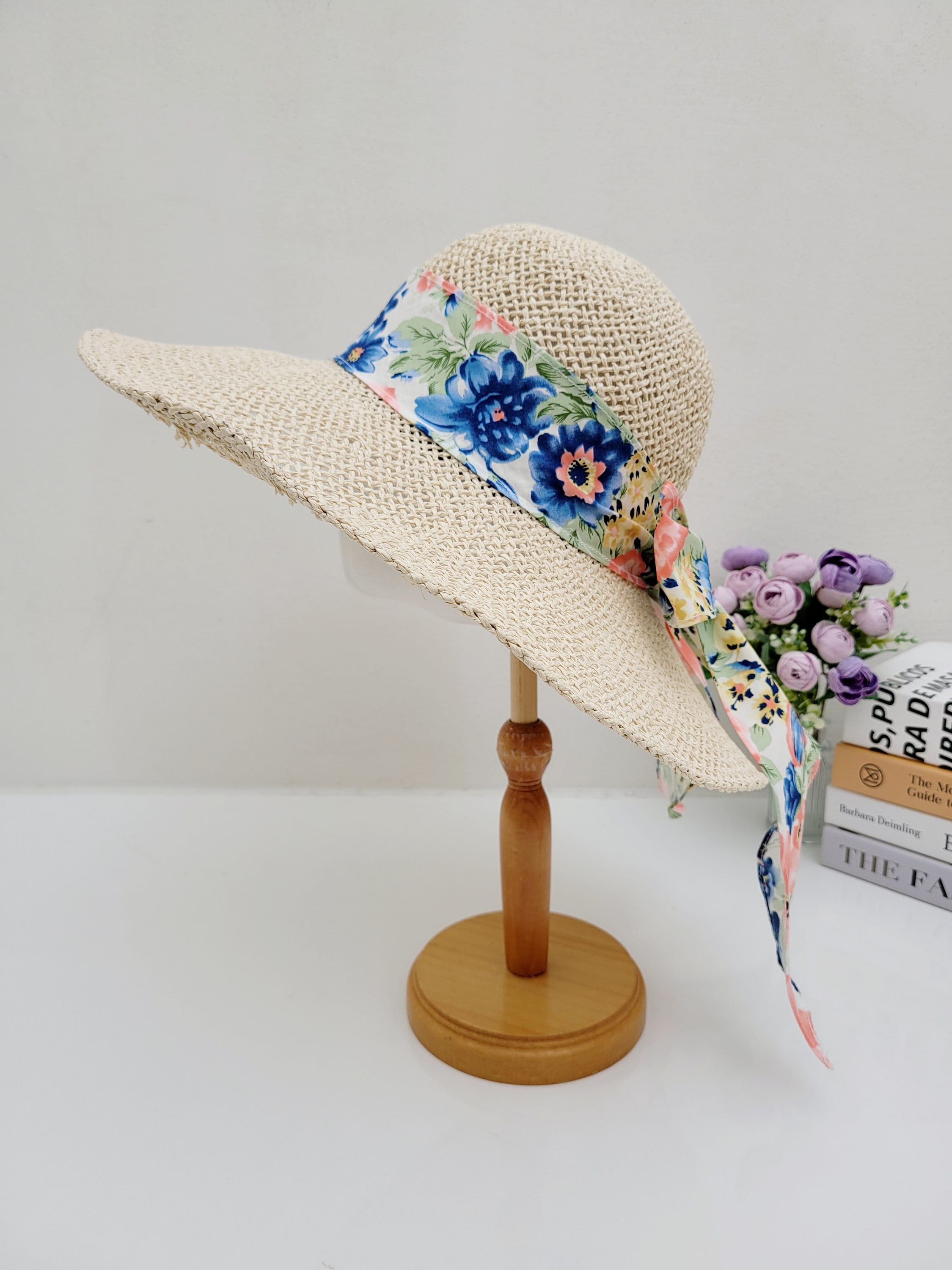 east hampton straw sun hat with a wide brim and a vibrant blue and pink floral ribbon detail on a wooden stand.