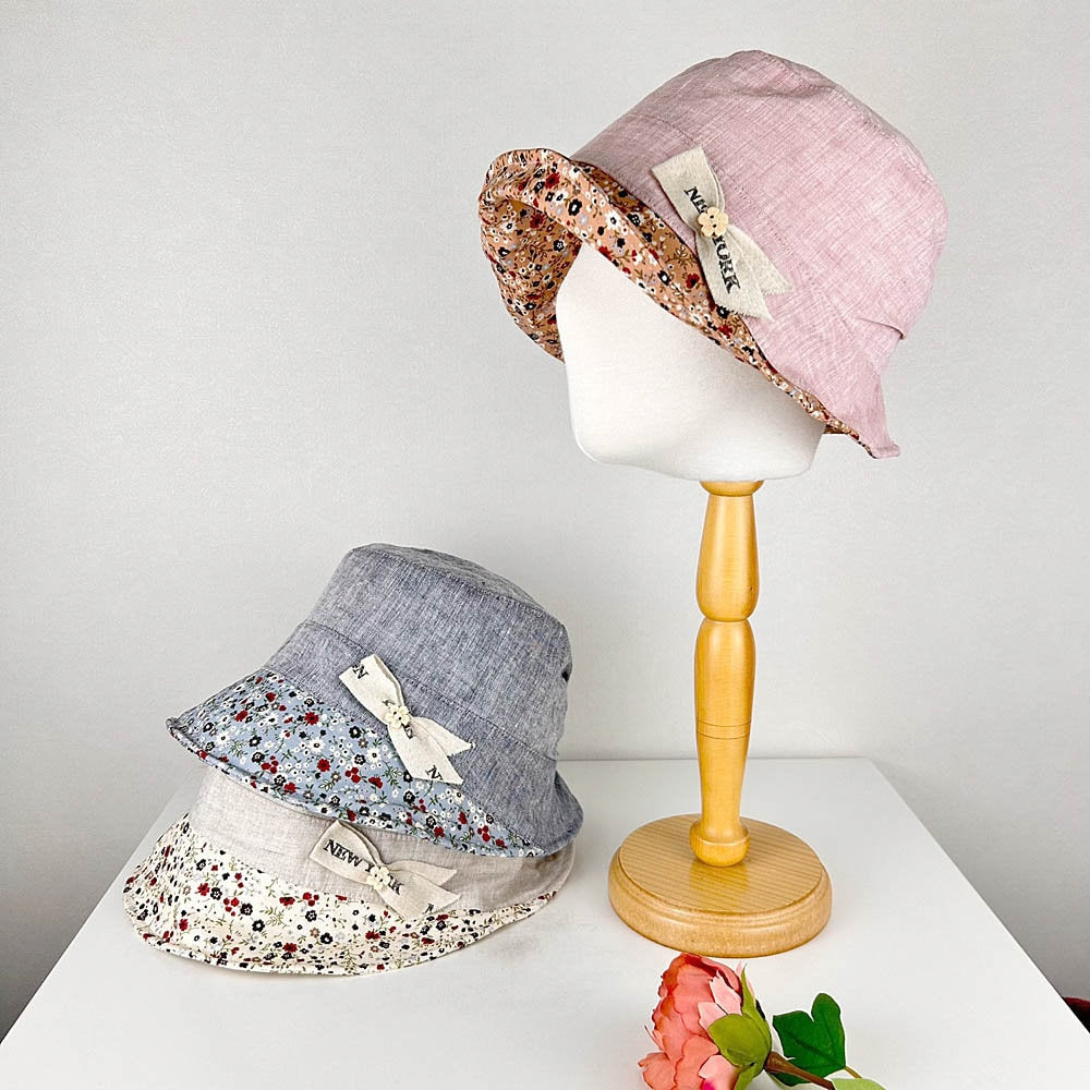 Olivia's Summer bucket hats: pink, gray, and beige linen with floral lining and "New York" ribbon detail, displayed on a table with a wooden stand.