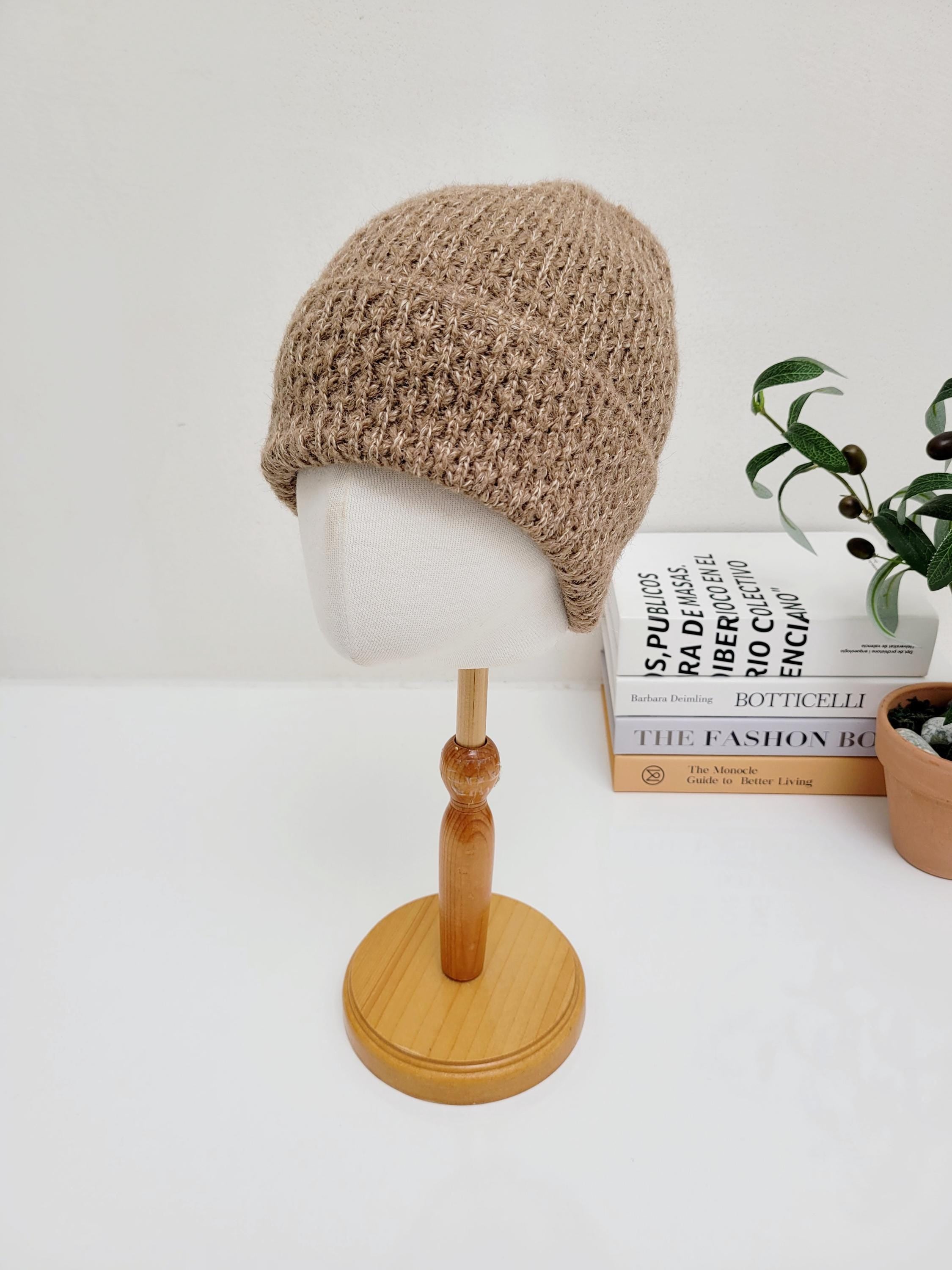 TAYLOR: Beige knit beanie on a mannequin head, displayed on a table with books and a small olive tree.