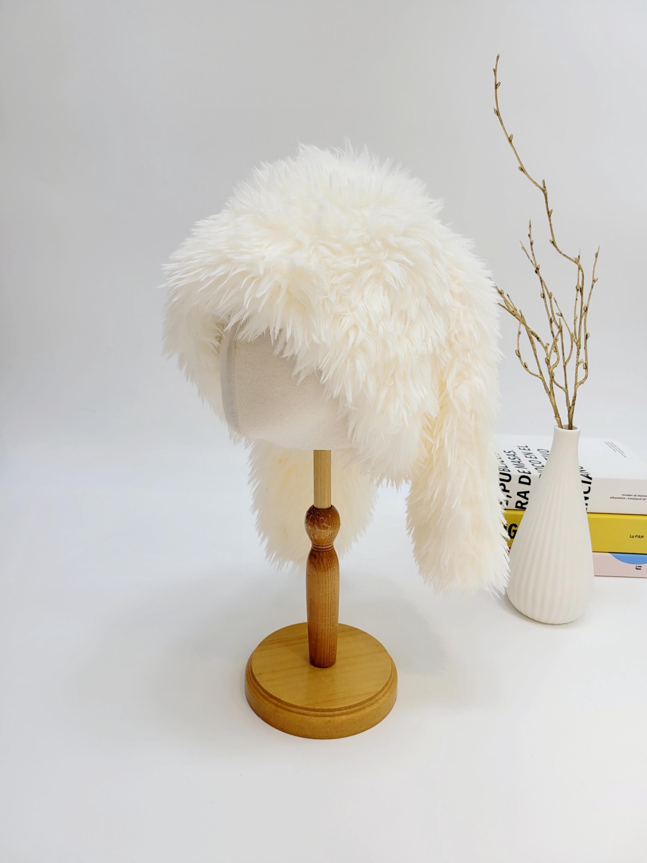 Fluffy white snow bunny hat with long ear flaps, displayed on a wooden stand. A cozy and stylish winter accessory.