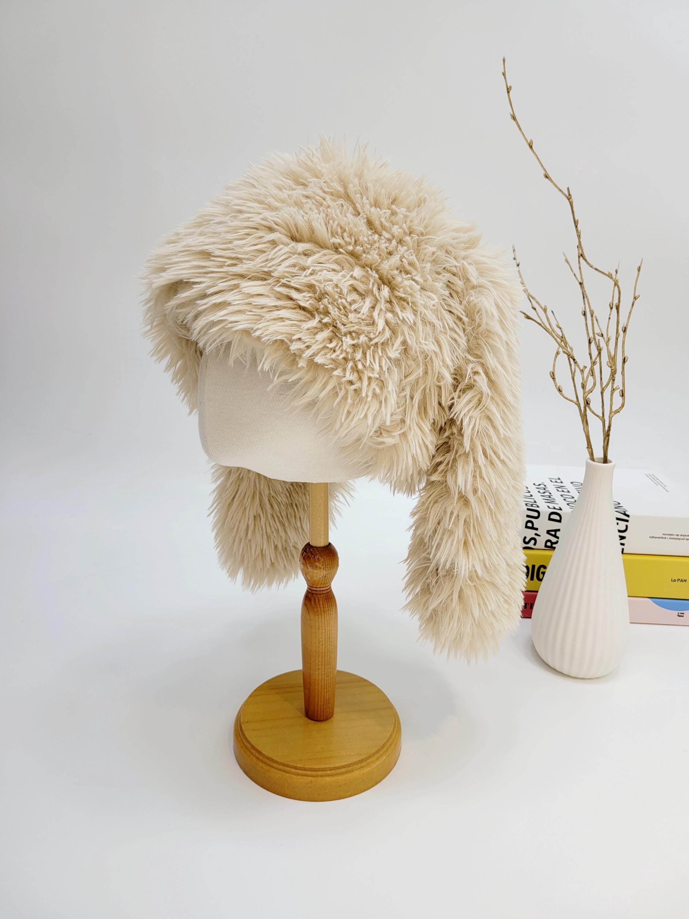 Creamy beige snow bunny hat. A soft, fuzzy hat with ear flaps displayed on a wooden stand. Perfect for winter warmth and style.