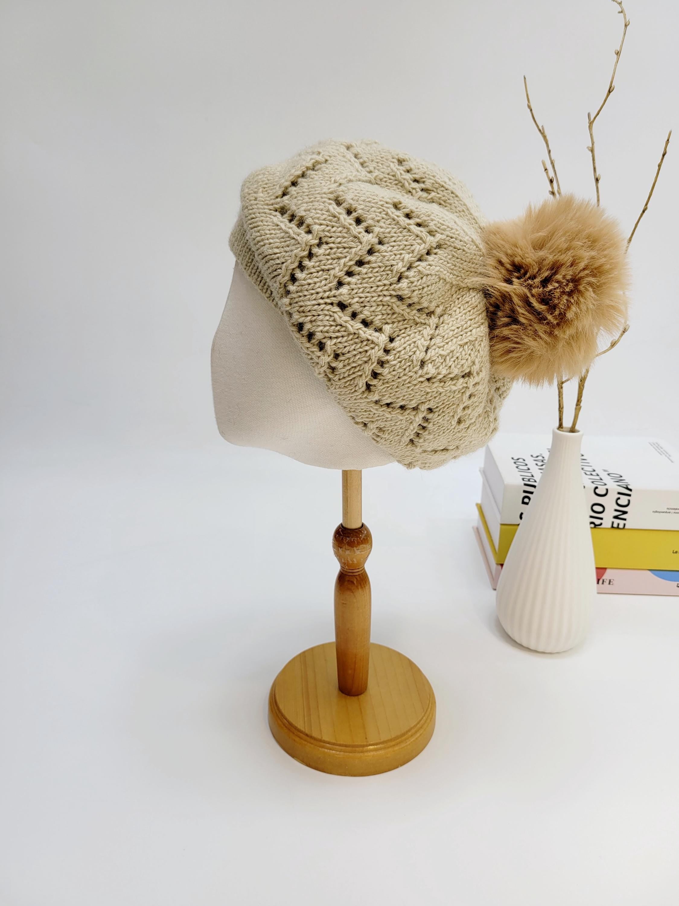 Emma in paris beige knit beret with openwork design and a fluffy pompom, displayed on a wooden stand.