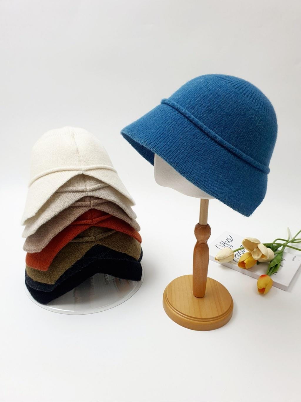 Stack of blair hats in cream, beige, brown, rust, black, and a single blue blair hat on a wooden stand with flowers.