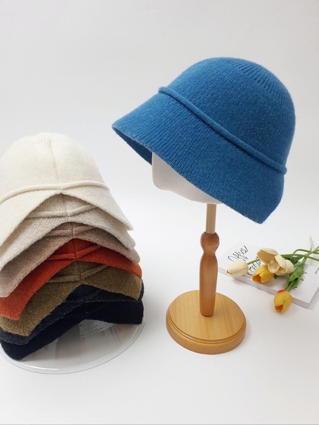 Stack of blair hats in cream, beige, orange, olive, and black. A blue blair hat is displayed on a wooden stand.