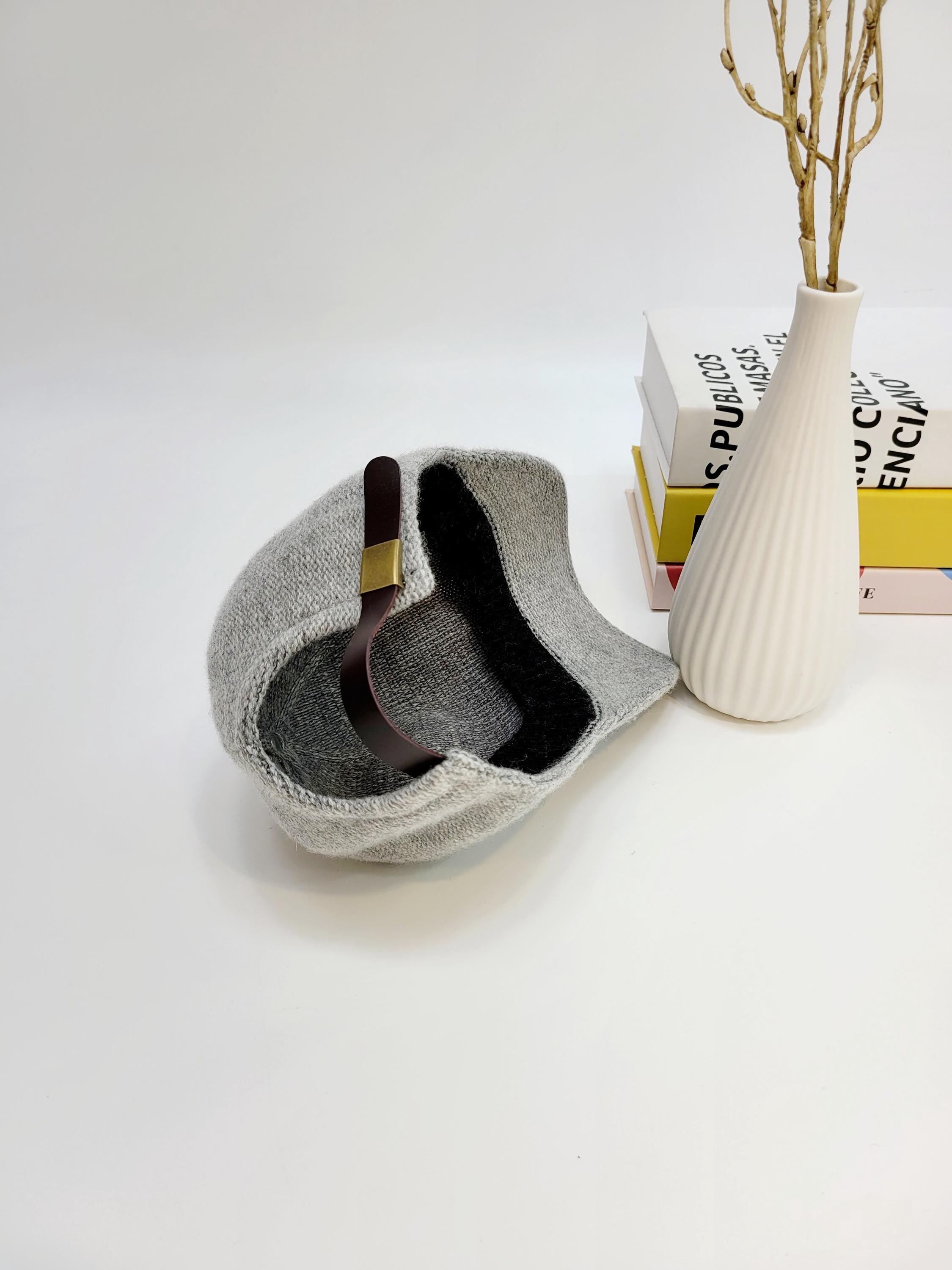 Gray wool cap with a brown leather strap detail, displayed next to books and a vase. The wool cap has a modern, stylish design.