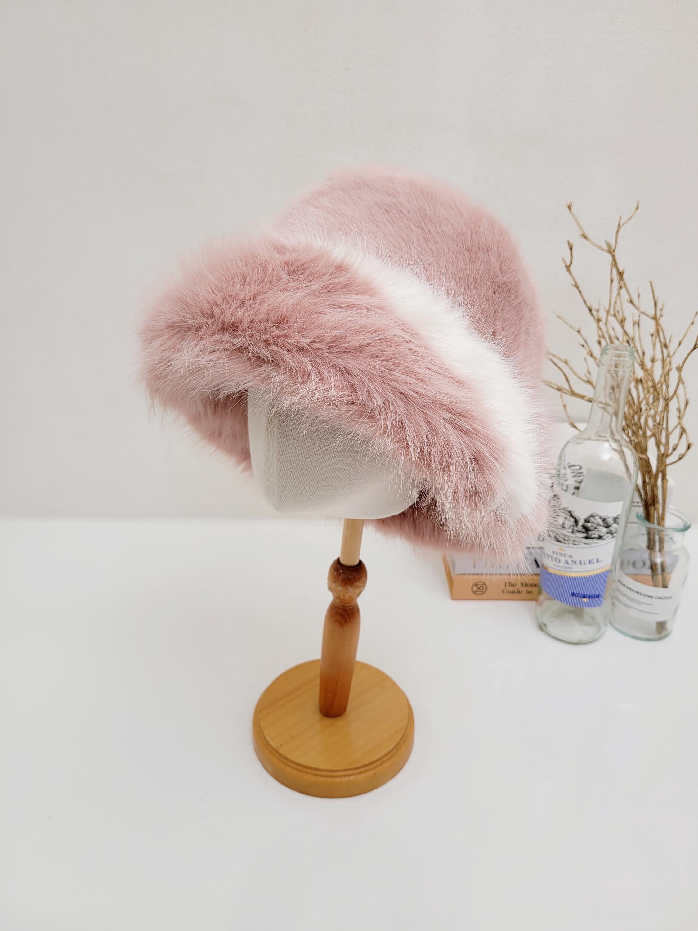 fluffy vegan fur hat in blush pink with a white band, displayed on a wooden stand
