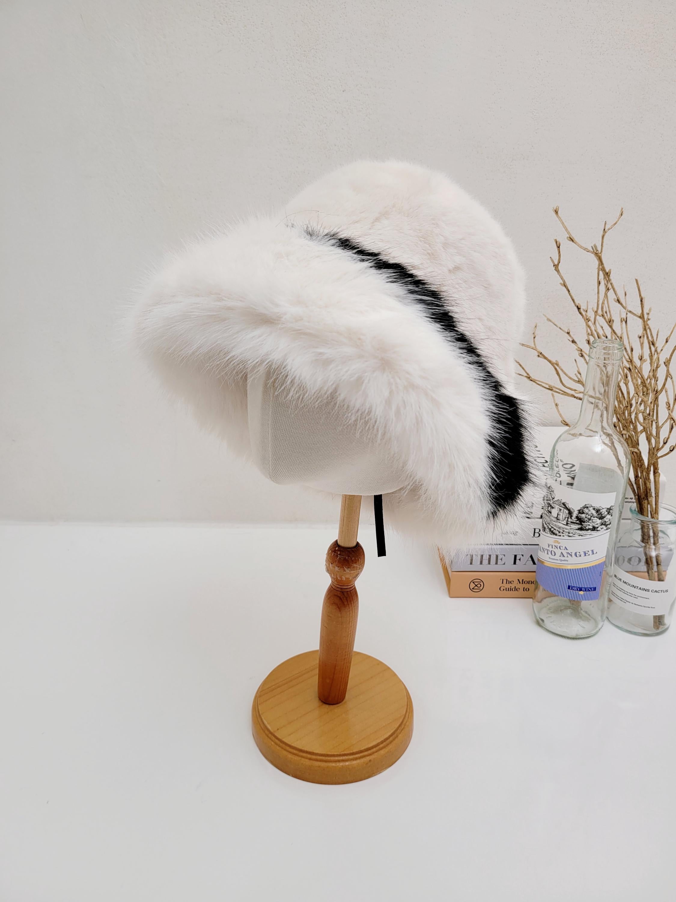 Fluffy vegan fur hat in white with a bold black stripe, displayed on a wooden mannequin stand. A stylish and cruelty-free headwear choice.