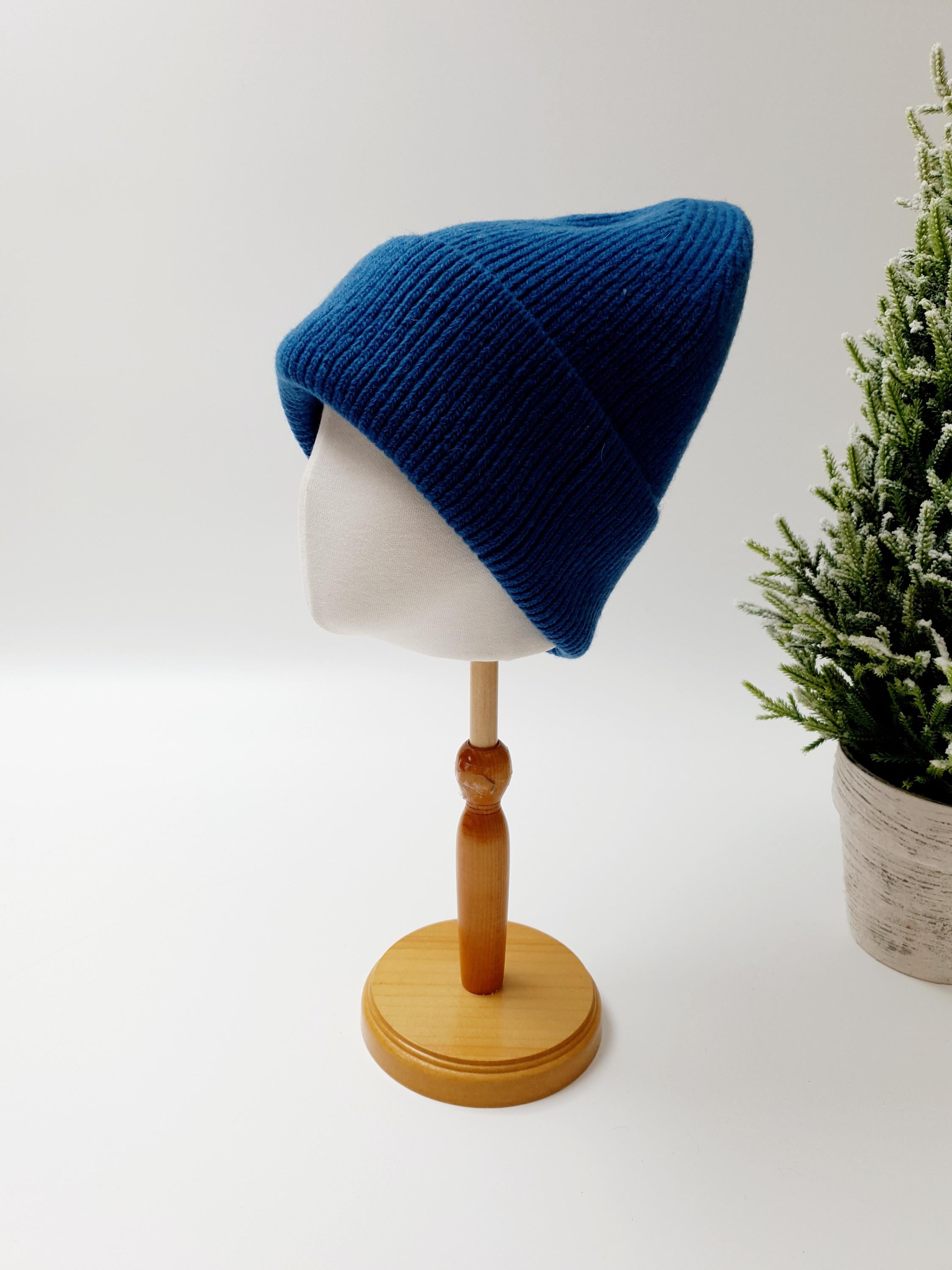 Dark blue ribbed beanie on a wooden stand next to a small potted evergreen tree, showcasing its knit texture and classic style