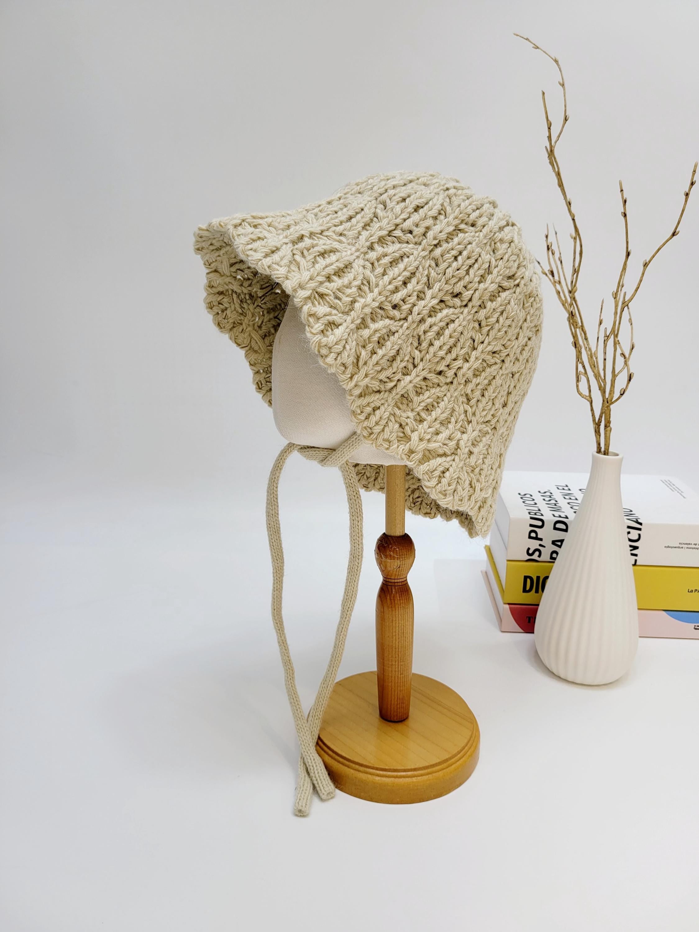Beige braided wool hat. Hand-knitted bonnet with a textured design and soft ties, displayed on a stand.