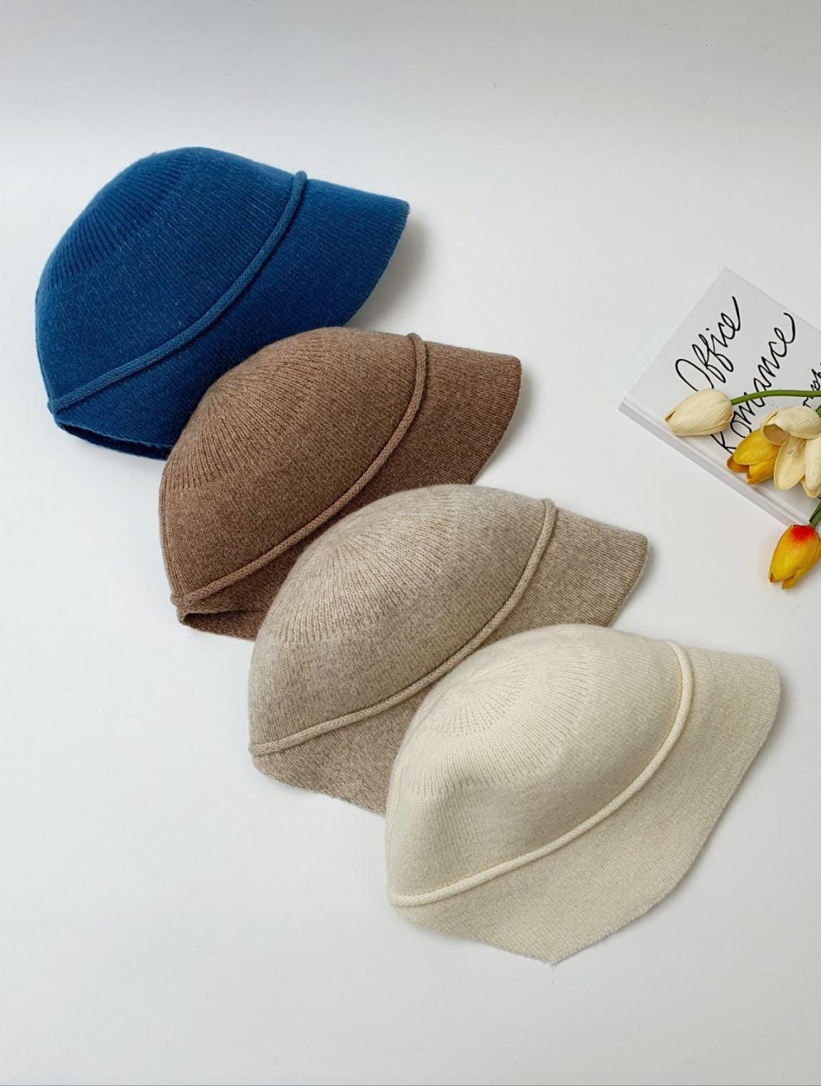 Four knitted blair hats in blue, brown, beige, and cream hues, stacked neatly beside a book and tulips
