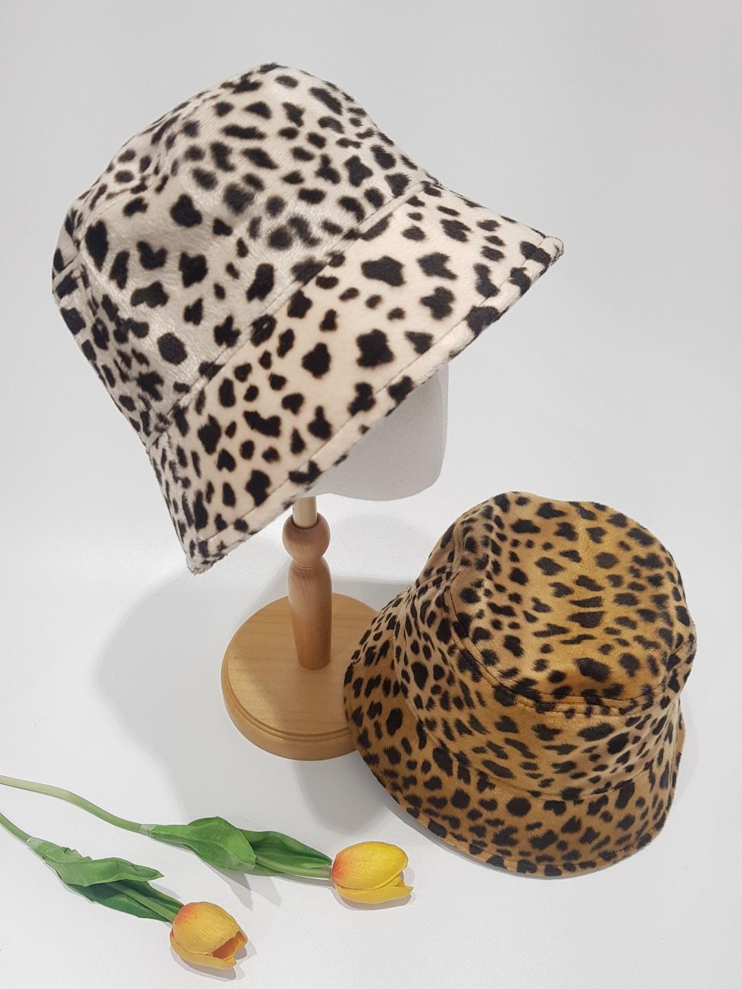 BIG CAT-BUCKET: Two bucket hats, one cream with black spots on a wooden stand, the other tan with black spots, with yellow tulips in the foreground.