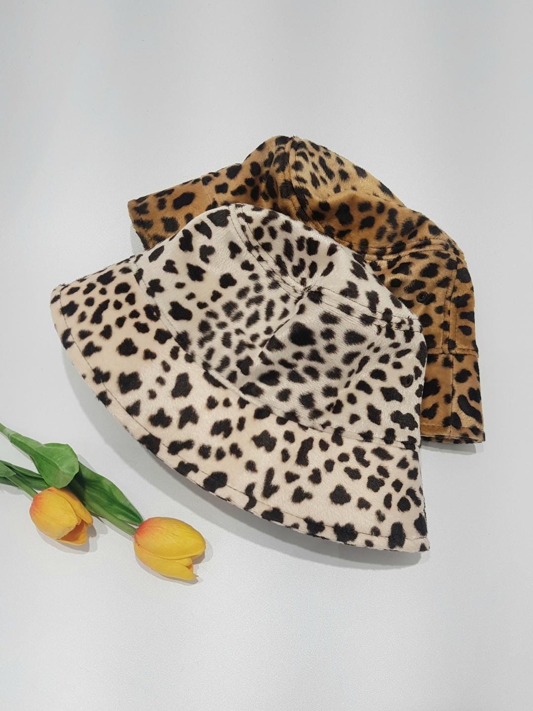 Two big cat-bucket hats: tan leopard print and cream Dalmatian print. Stylish animal print bucket hats next to yellow tulips.