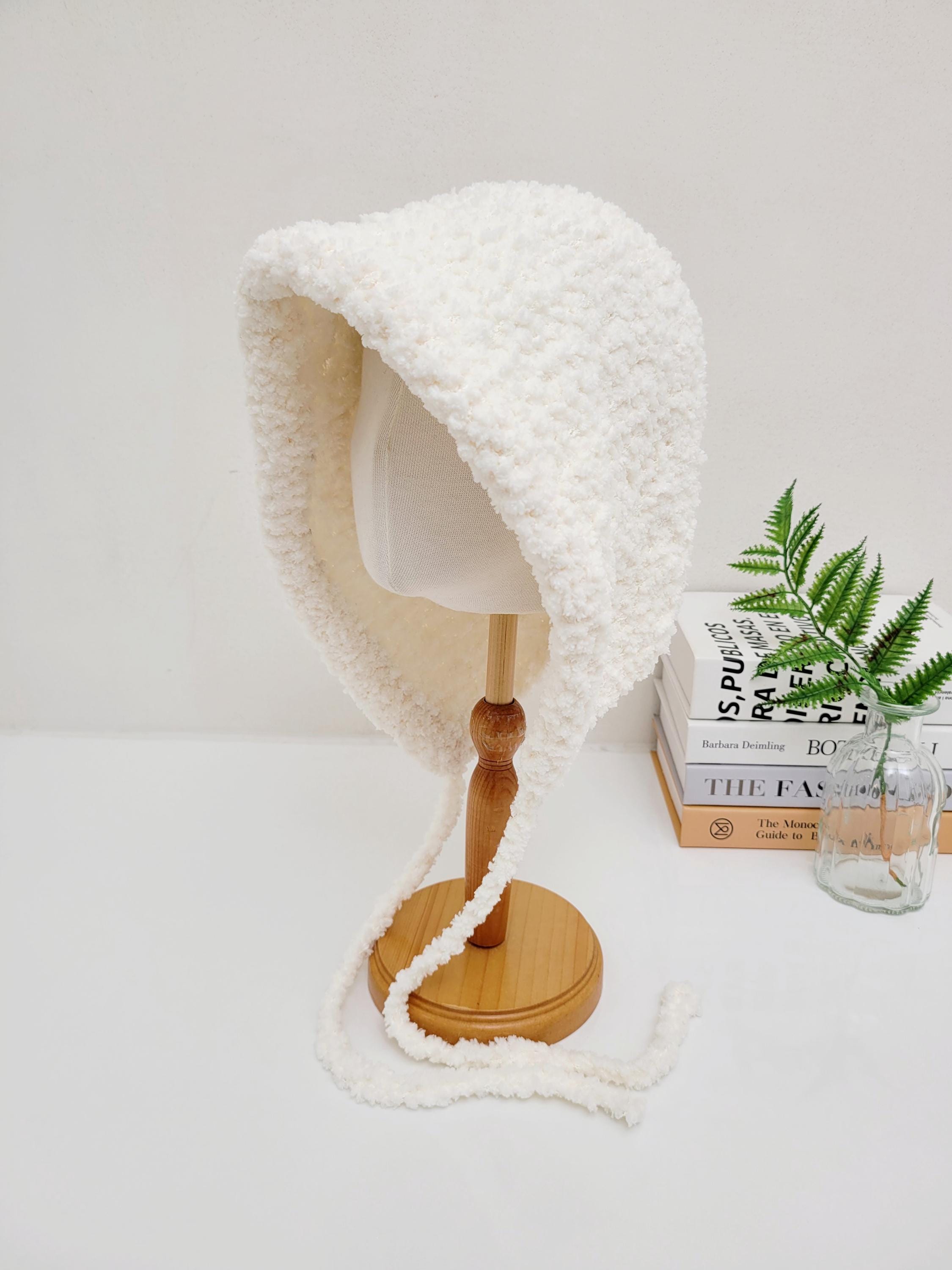 little riding hood - sherpa bonnet: Soft, textured white sherpa bonnet with tie closures, displayed on a wooden stand.