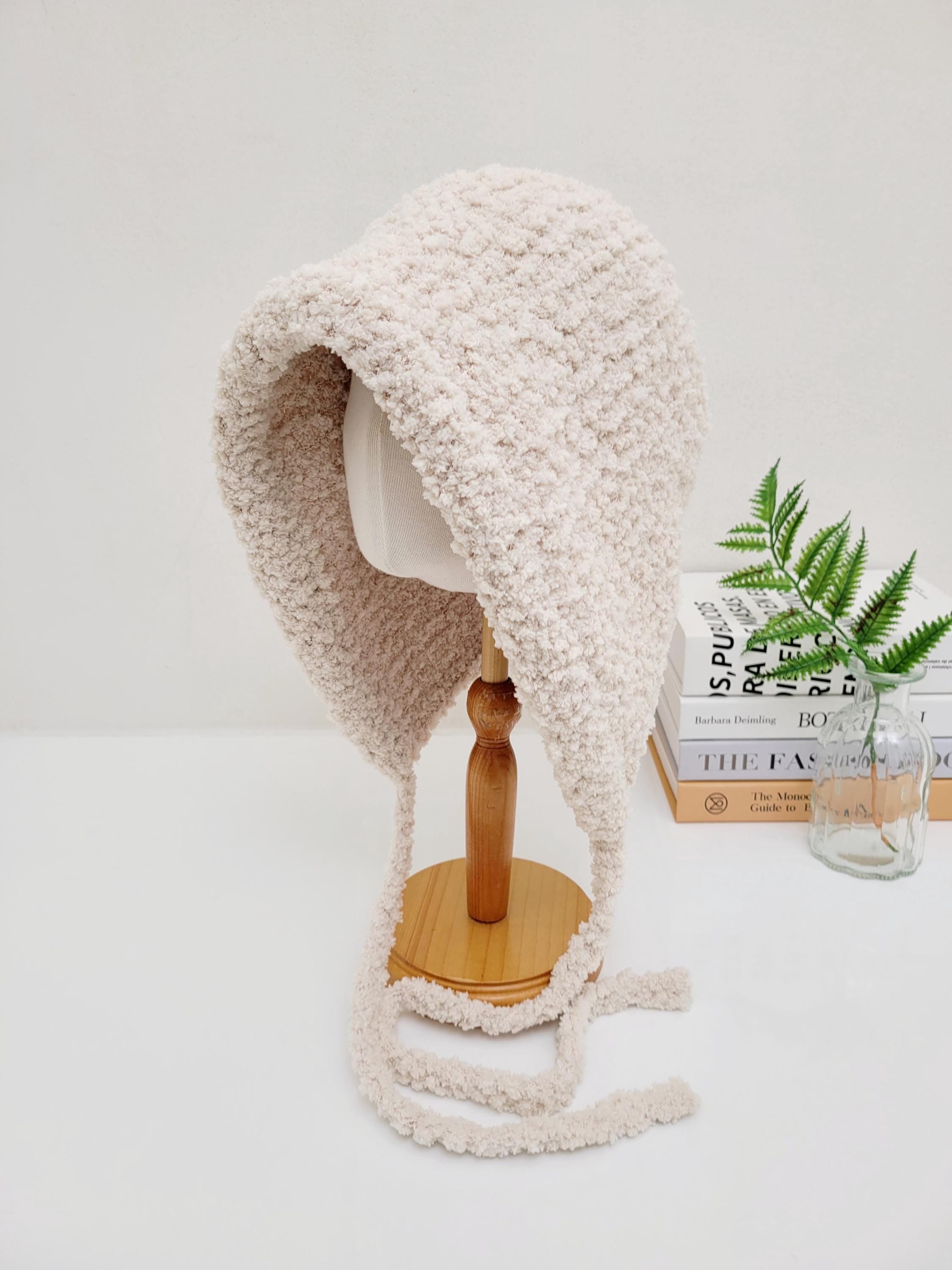 little riding hood - sherpa bonnet in soft beige sherpa fleece with tie strings, displayed on a wooden stand.