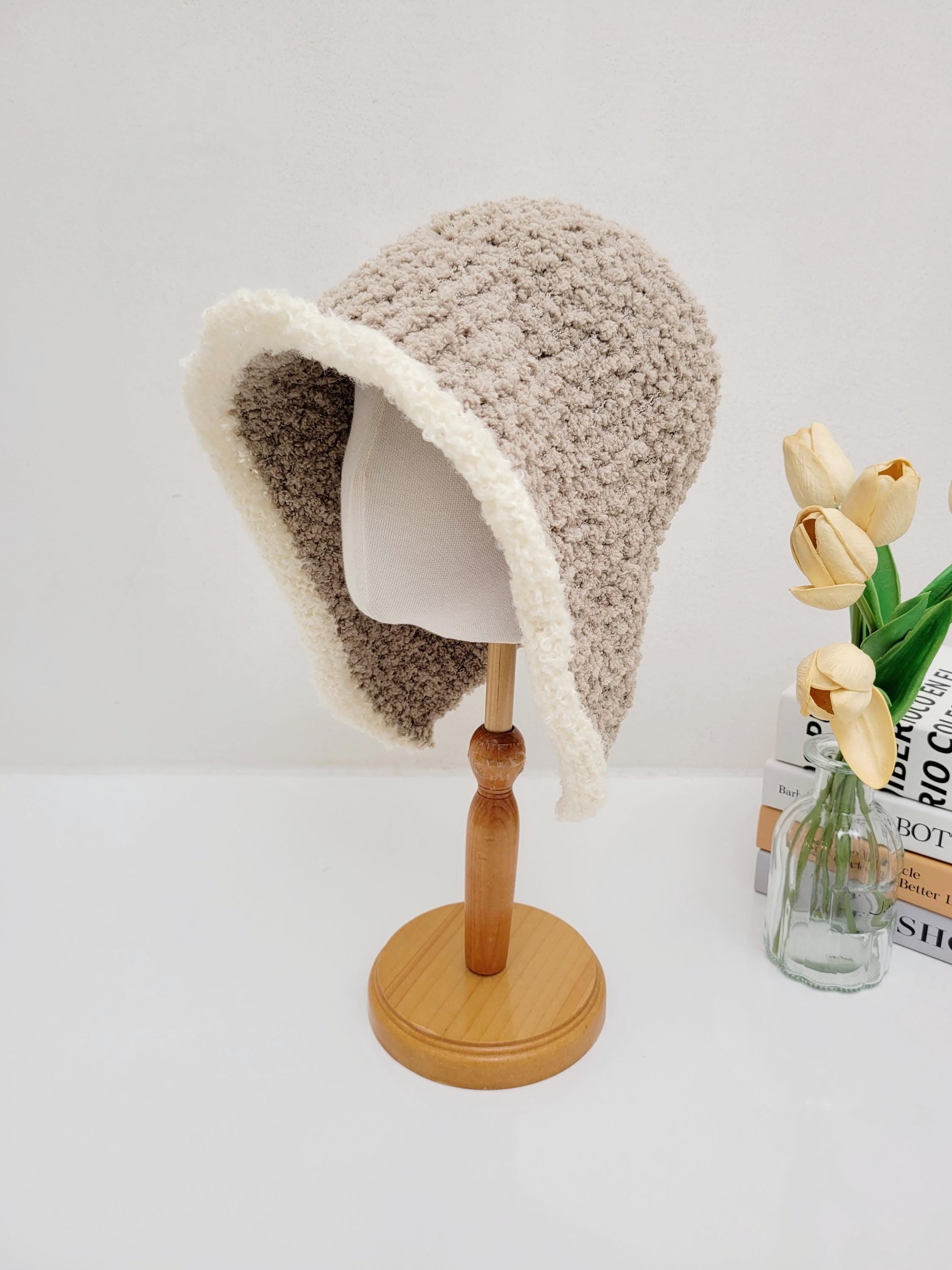 Beige wool hooded hat with a soft, white fluffy brim, displayed on a wooden stand. The textured knit hat offers warmth and style.