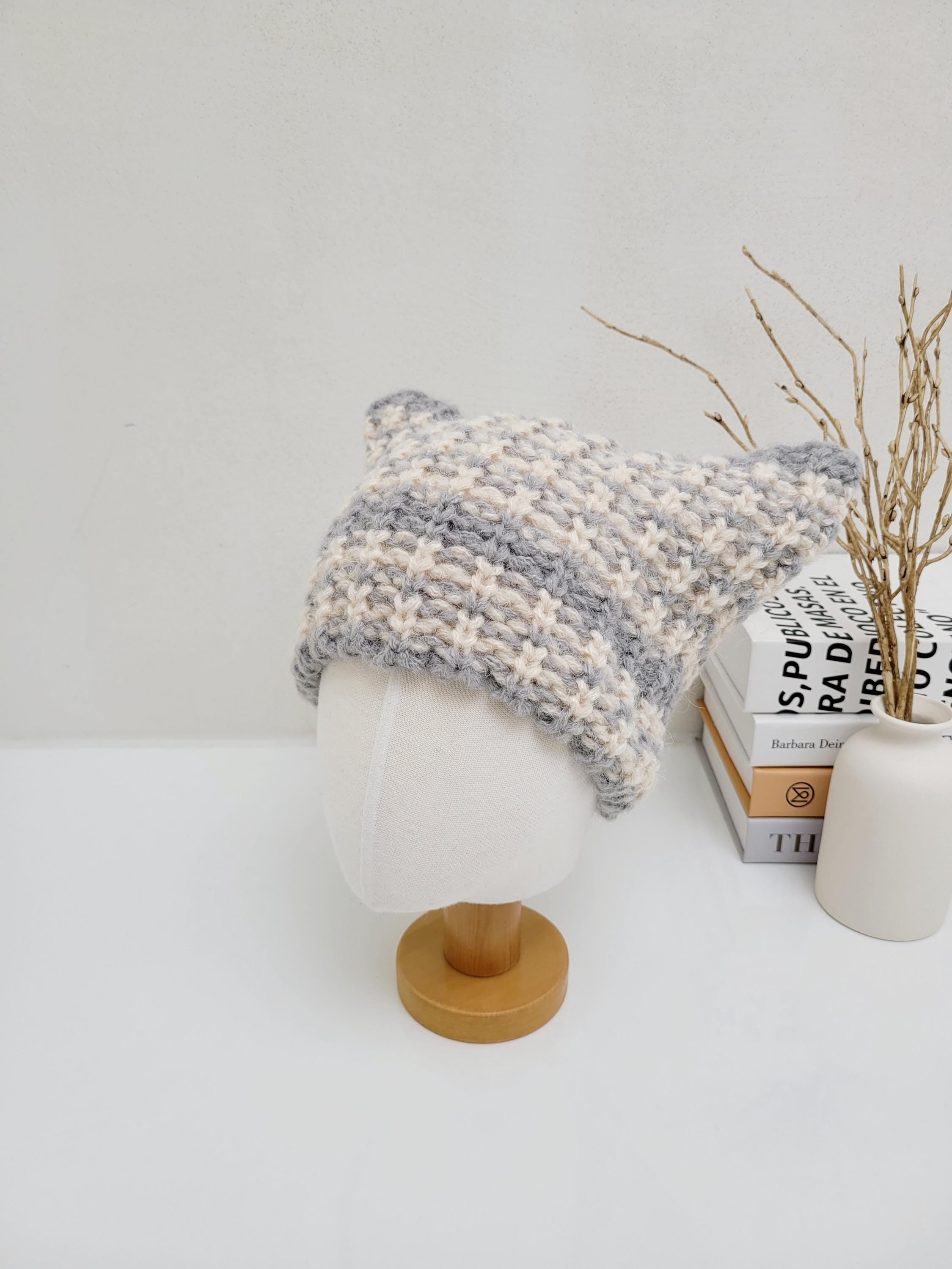 Grey and white knitted cat ear hat on a mannequin head, featuring pointed ears and a textured pattern.