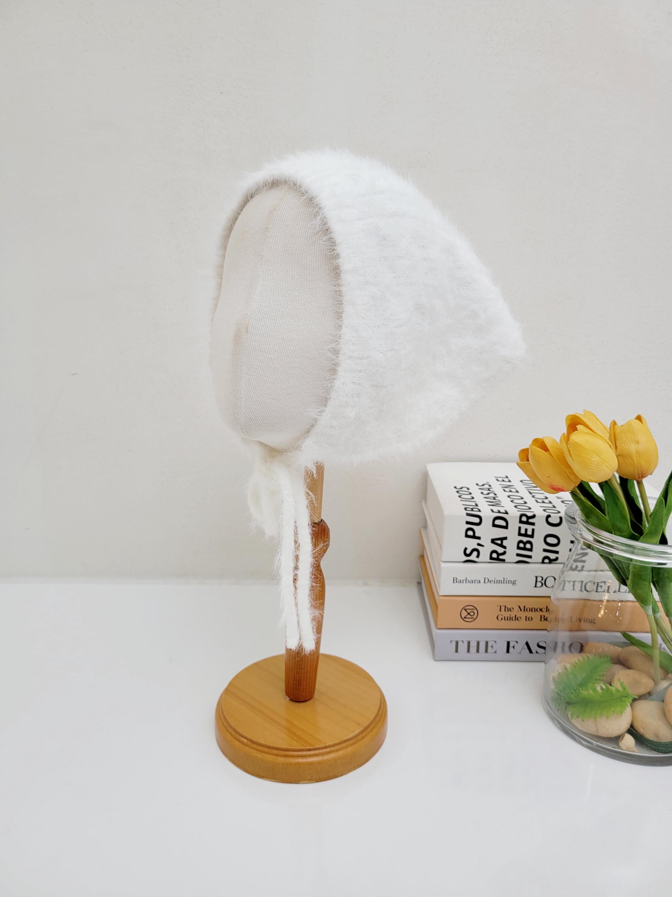White, fuzzy "little riding hood" baby bonnet with soft ties, displayed on a wooden stand.