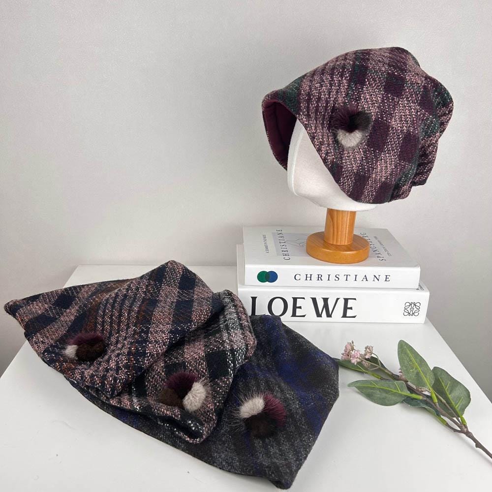 Checkered slouch hat and neck warmer set. The hat is displayed on a mannequin head, while the neck warmer is folded on the table. Both are made with a checkered pattern fabric and decorated with pom-poms.