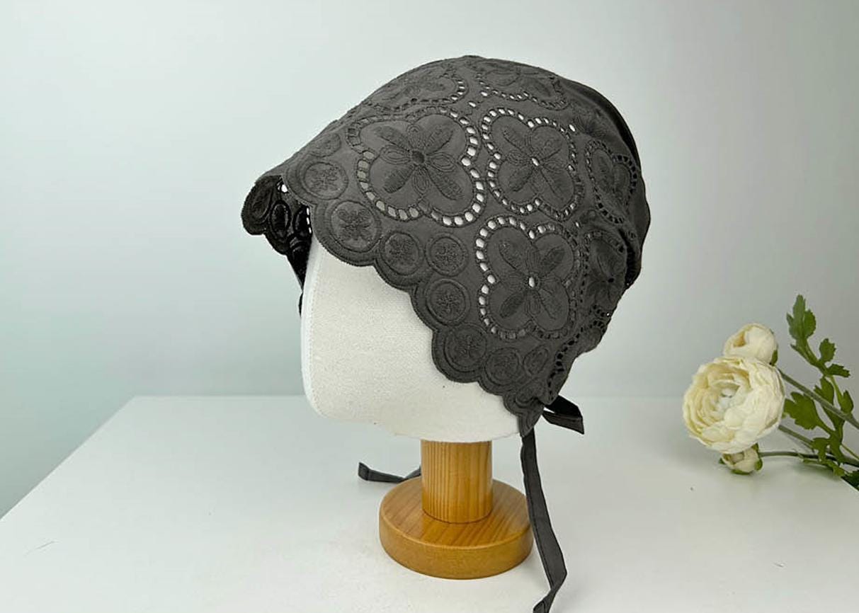 Gray eyelet beanie on display. This bonnet features a delicate floral cut-out pattern and ribbon ties.