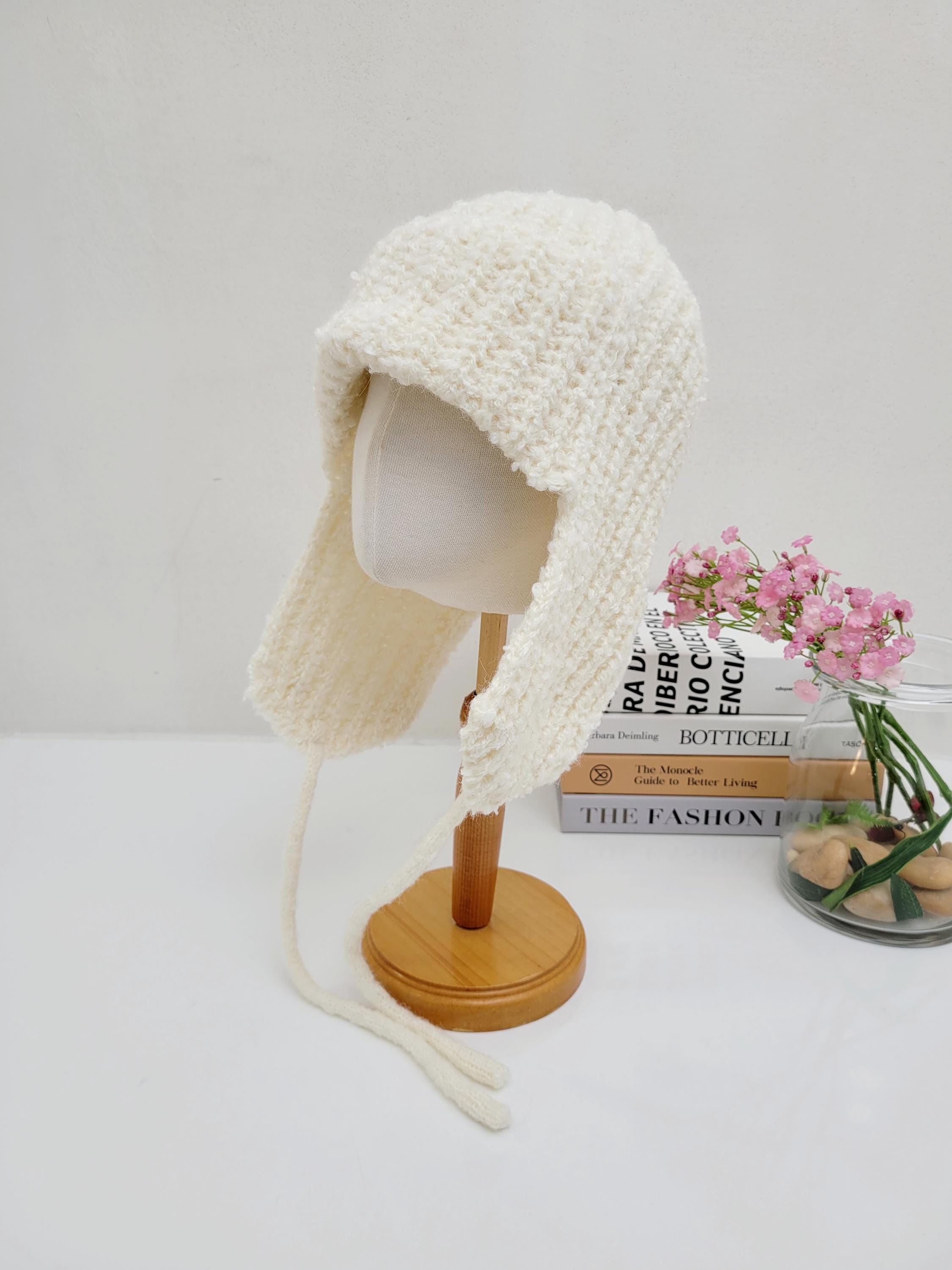 Cream knitted wool earflap hat with long braided ties, displayed on a wooden stand. Cozy and stylish winter headwear.