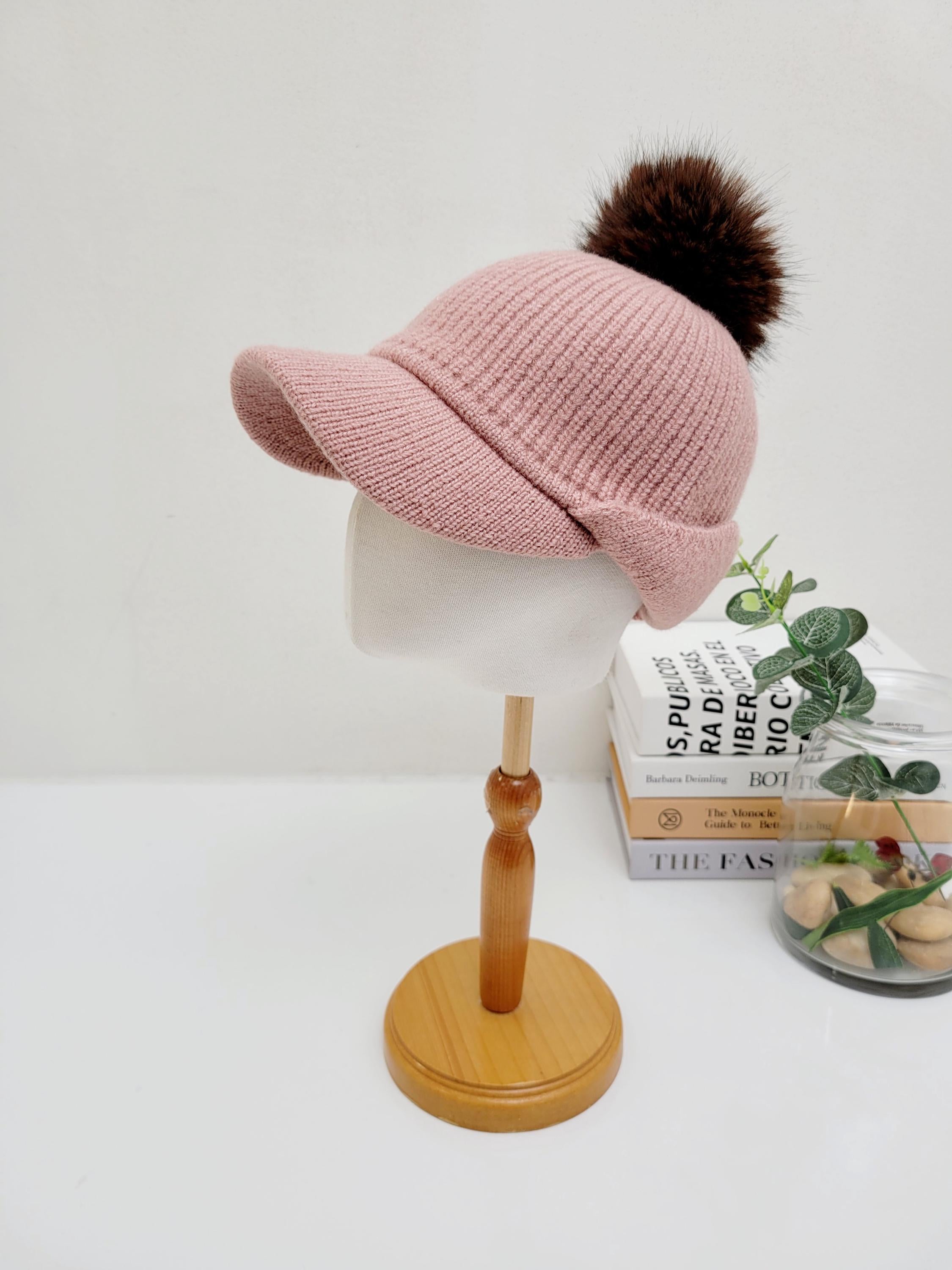 Pink ribbed cap - pom with a brim and fluffy brown pompom, displayed on a stand.
