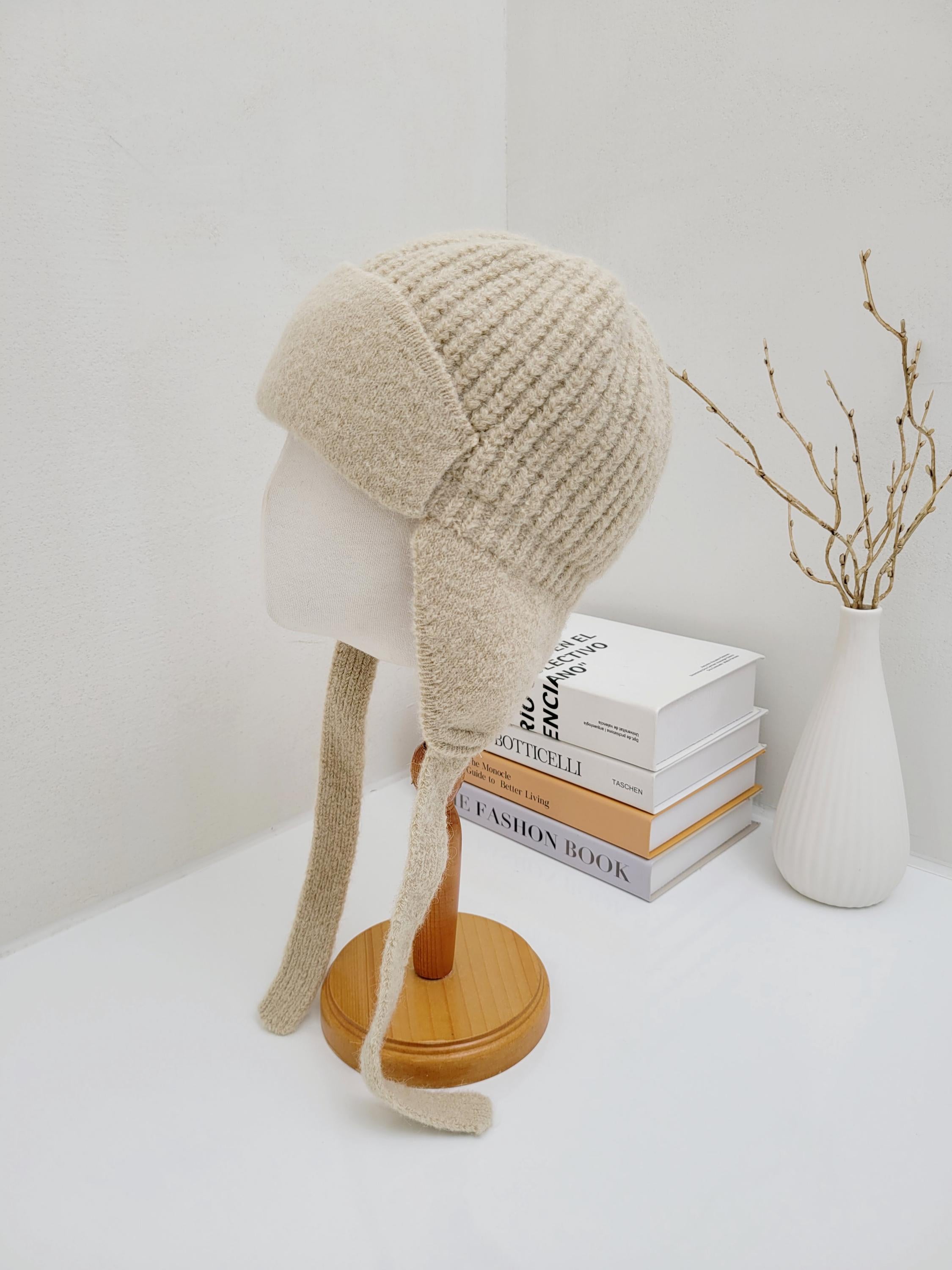 AMELIA: Beige knit hat on a mannequin, featuring ear flaps and tie closures for warmth & style.