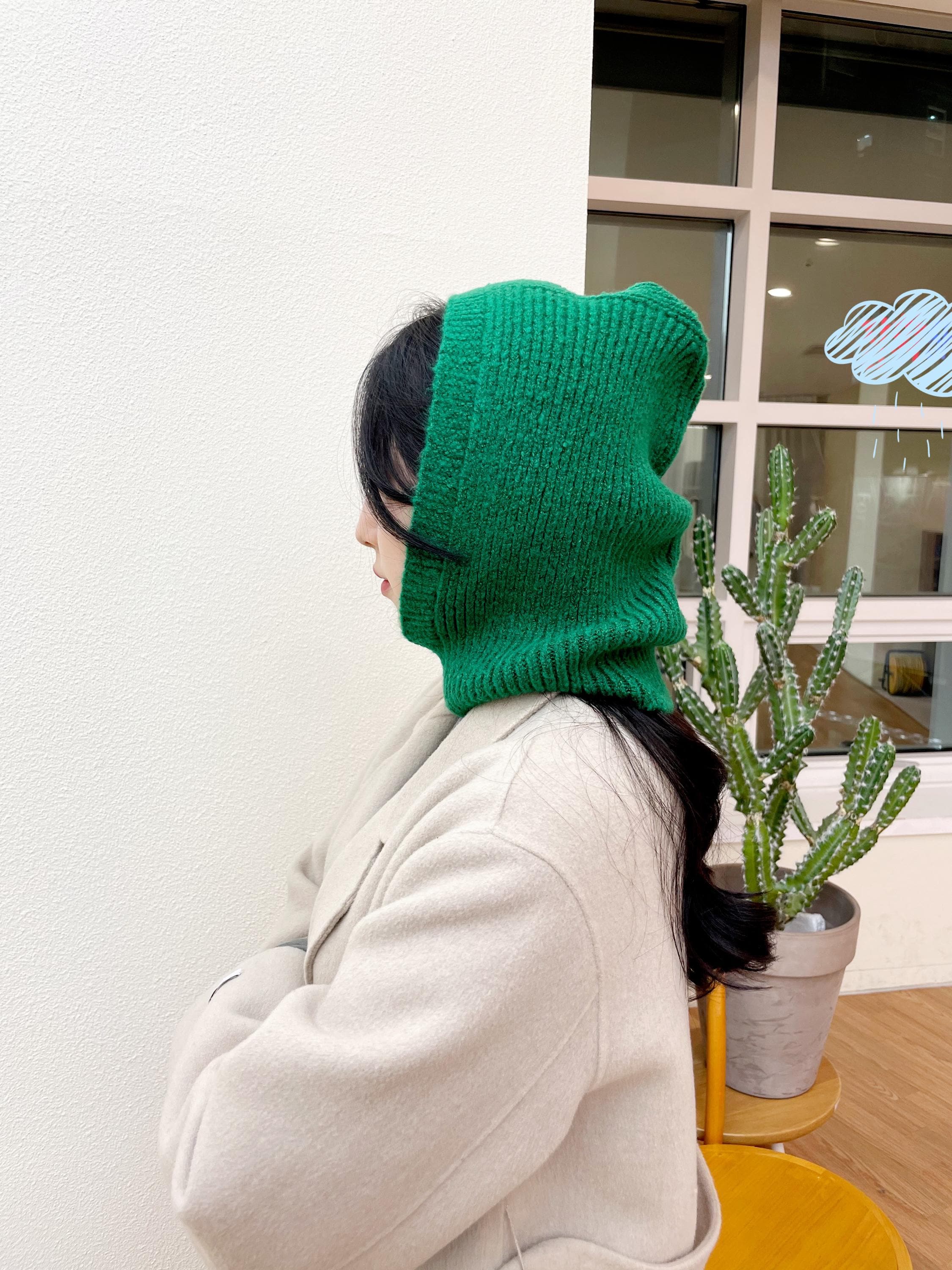 Green knitted little riding hood - balaclava on a woman, side view. She wears a cream coat, standing against a white wall.