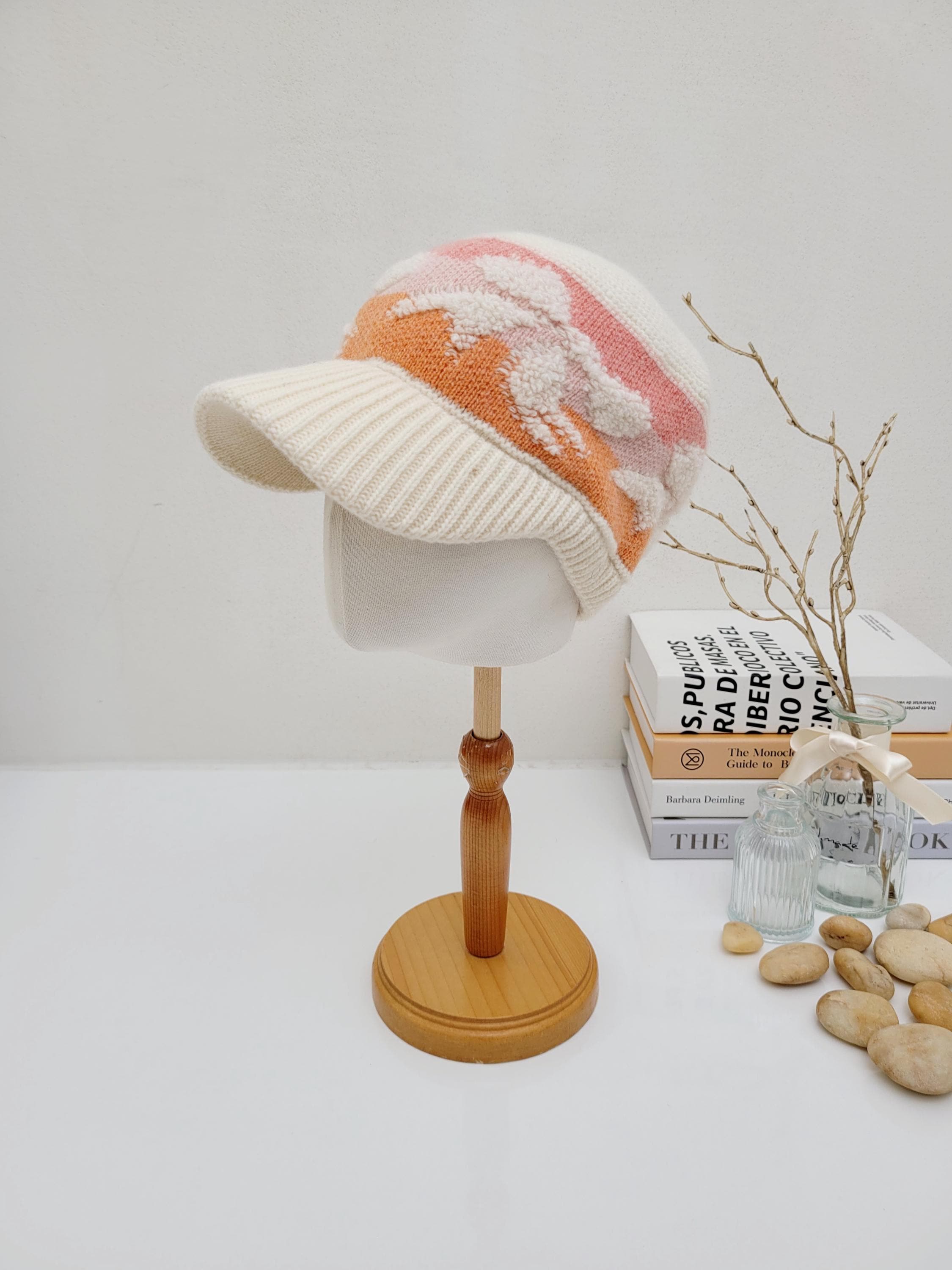 Cream knitted wool cap with a visor, featuring a pink and orange floral design. The earmuff knitted wool cap is displayed on a wooden stand.