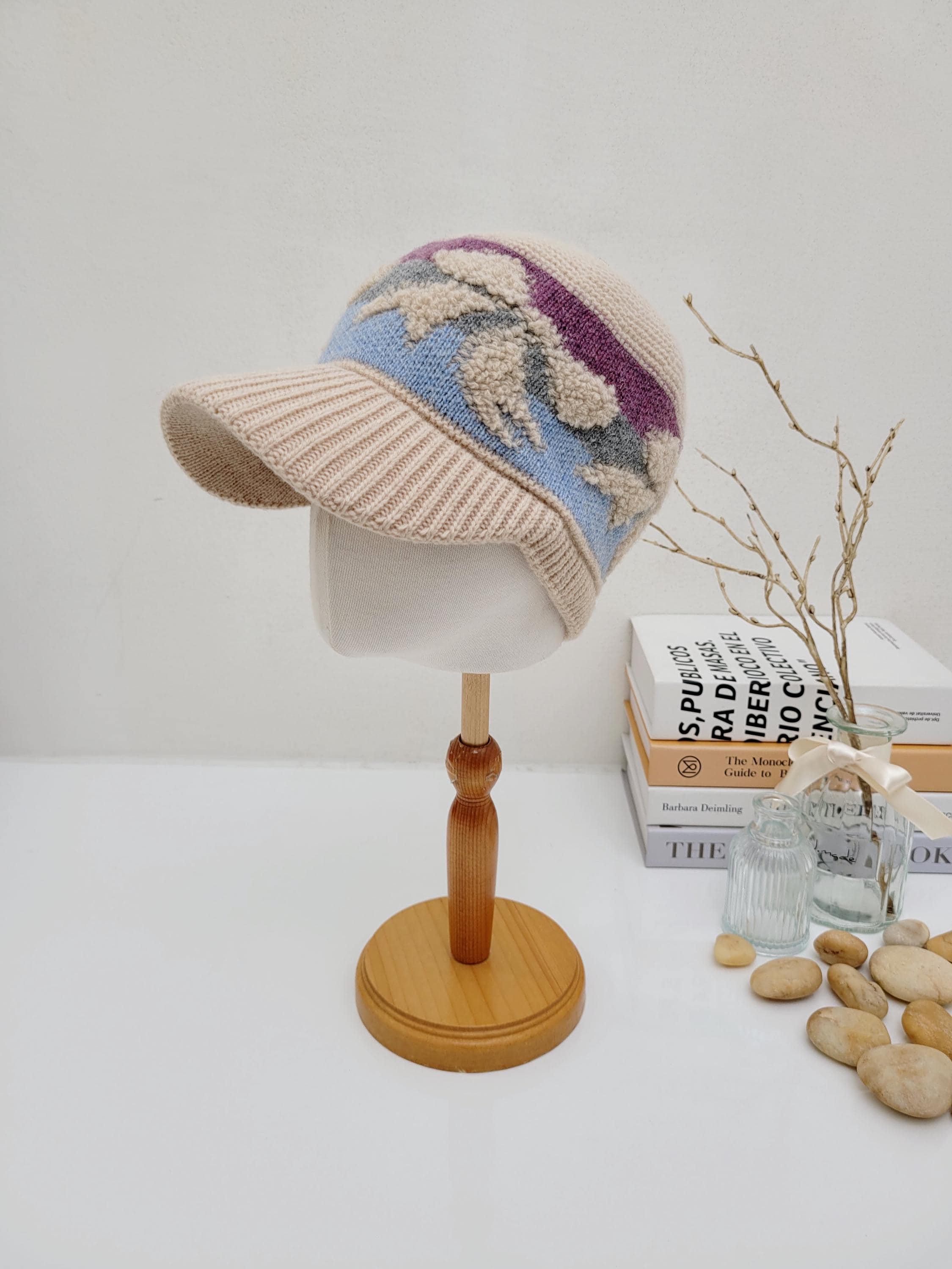 EARMUFF KNITTED WOOL CAP: Beige knitted wool cap with a visor, featuring blue, purple, and textured white floral accents. Displayed on a mannequin head.